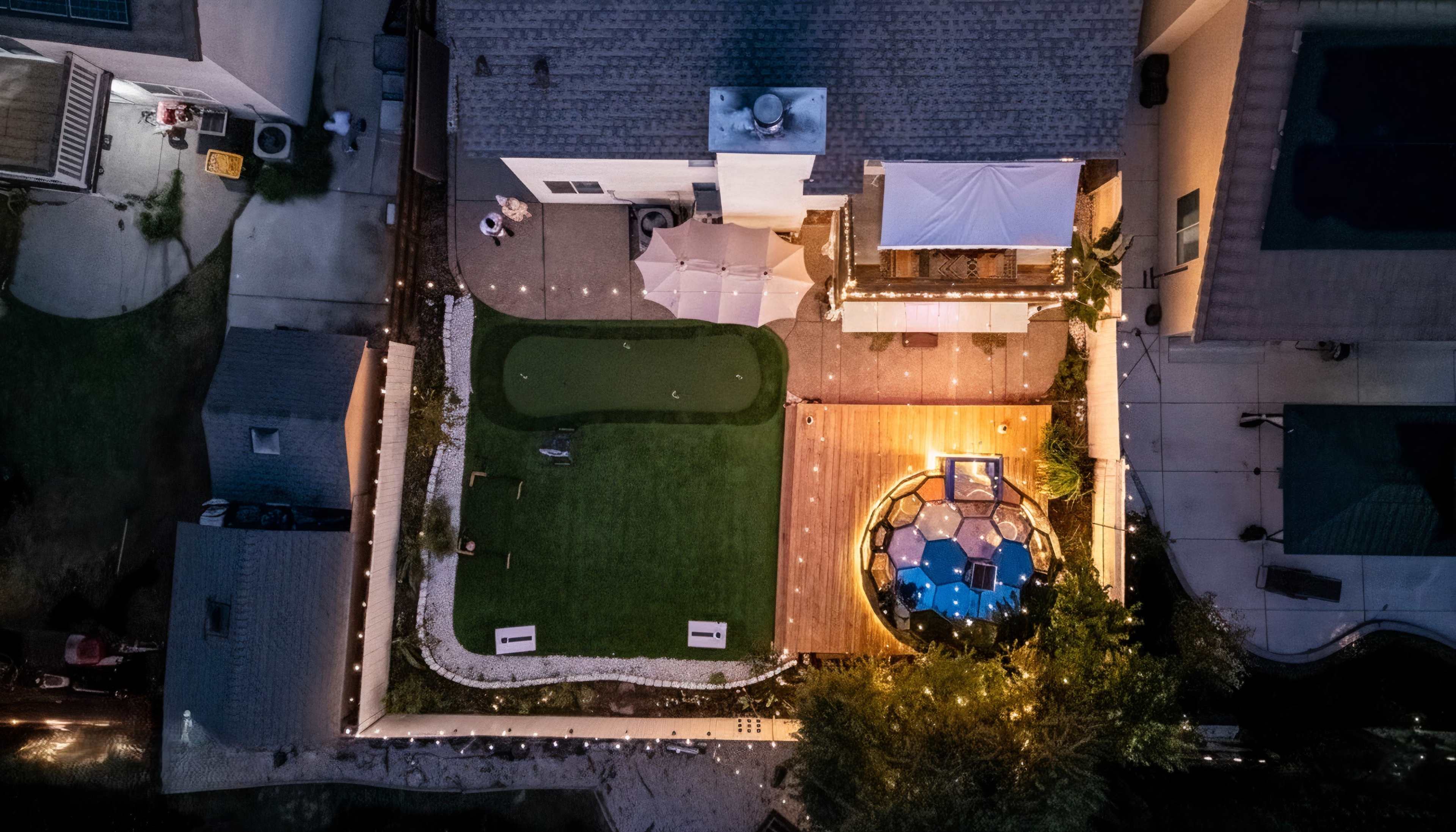 The image shows an aerial view of a backyard featuring a putting green, seating area with a table, a hot tub, and various outdoor lighting.