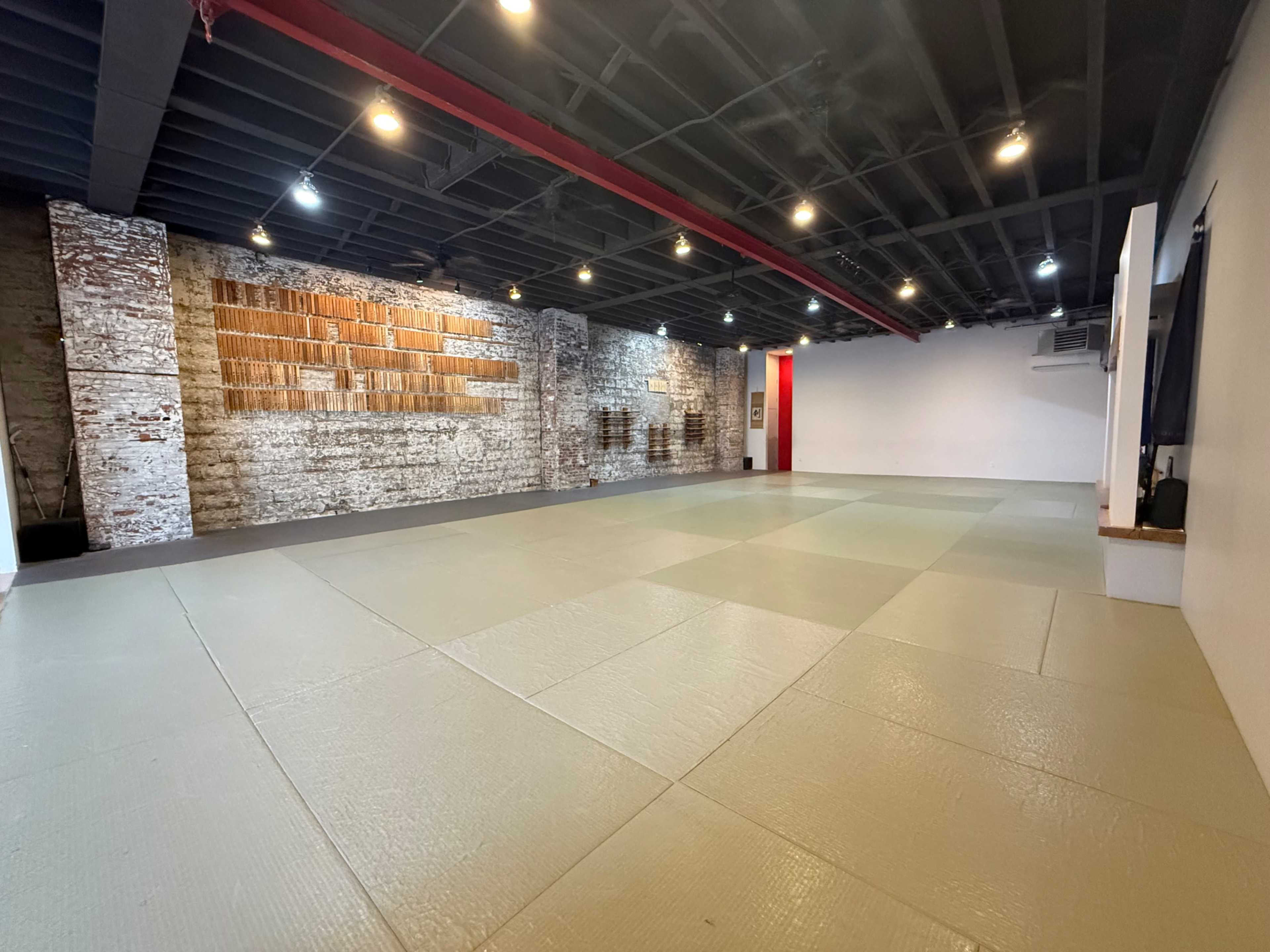 The image shows an empty dojo space with smooth, light-colored mats and exposed brick walls.