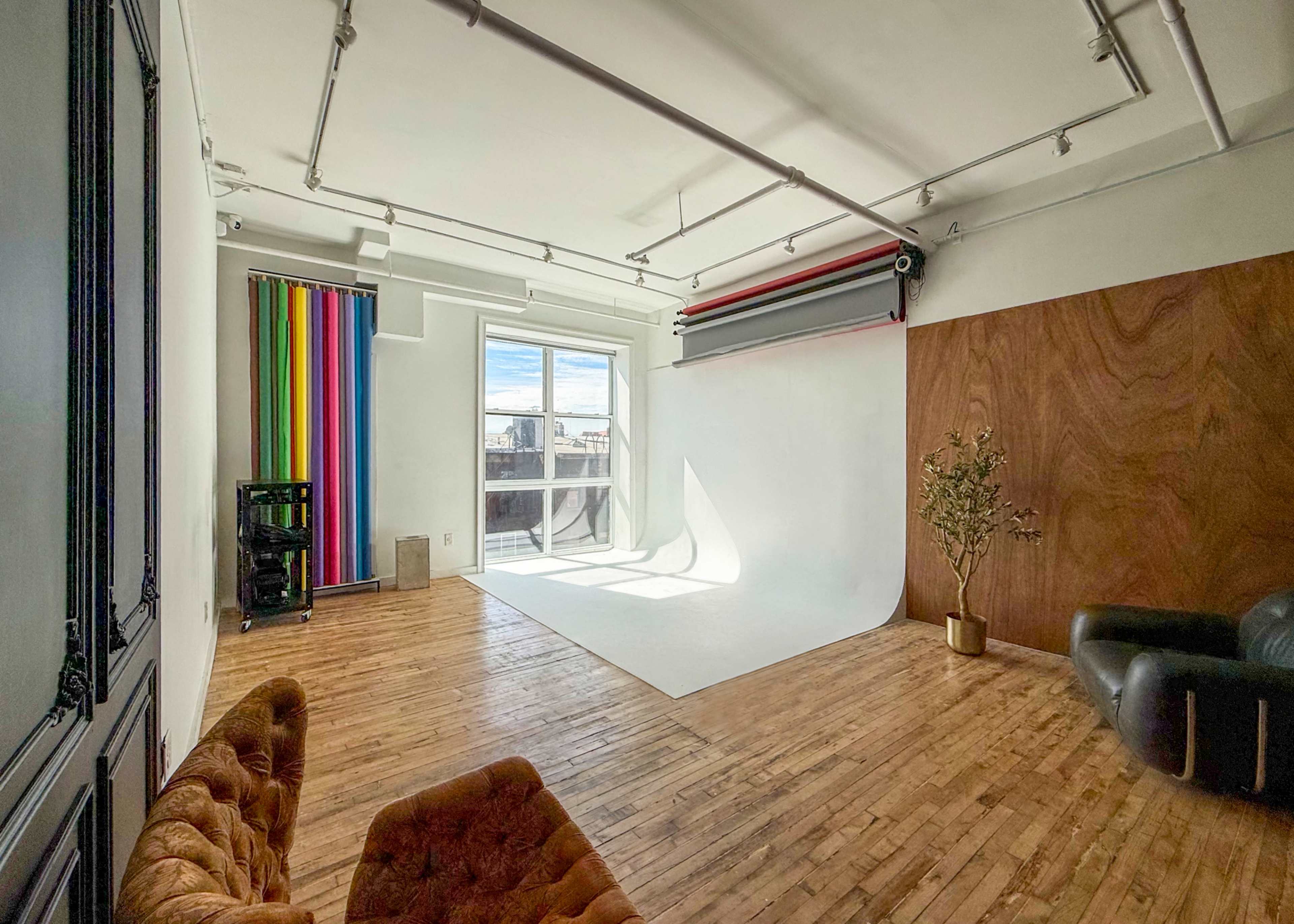 The image shows a spacious, well-lit room with wooden flooring, a white backdrop, and colorful curtains on one side.
