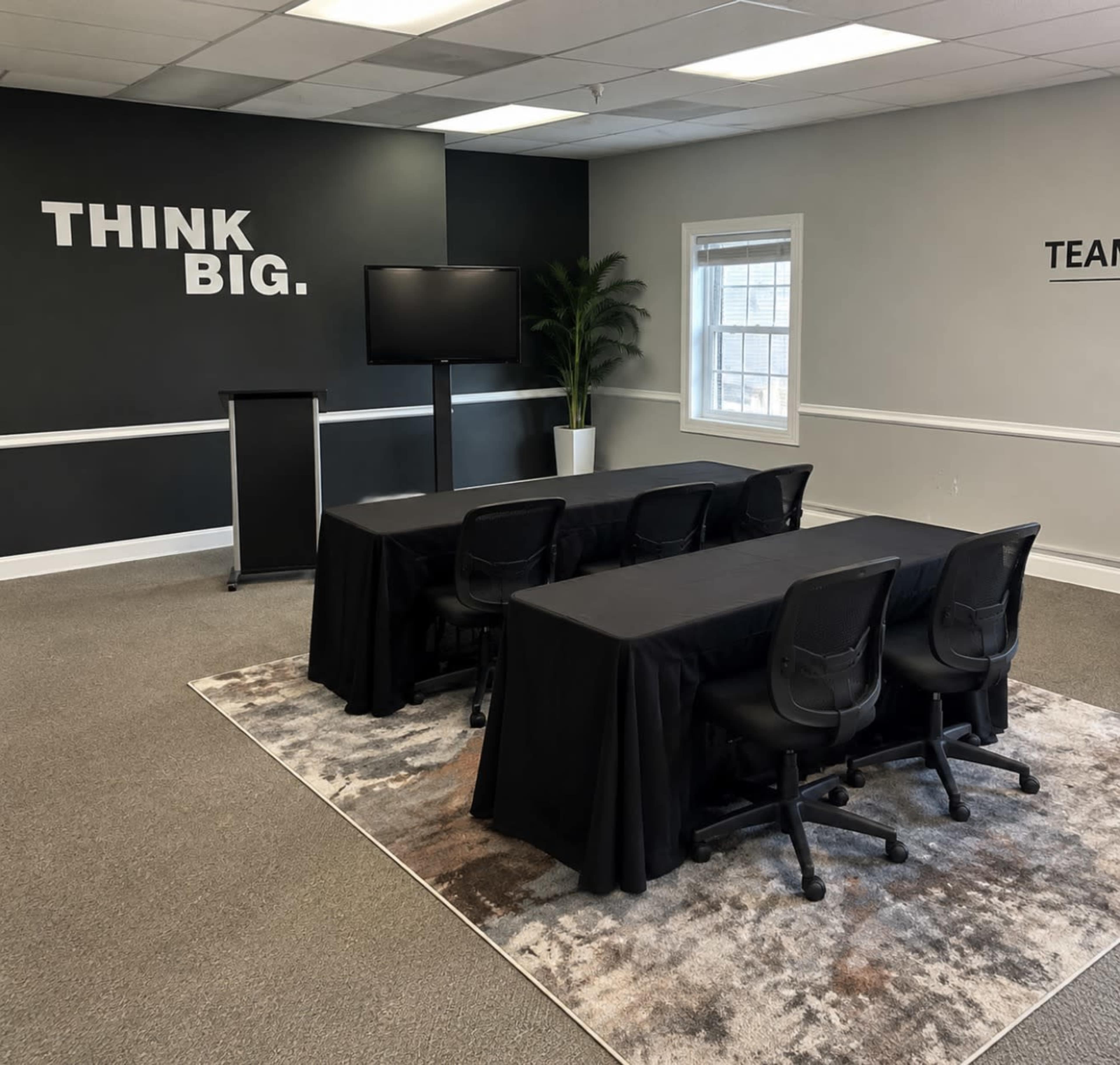 A meeting room features a black table setup with chairs, a television on a stand, and motivational wall decor.
