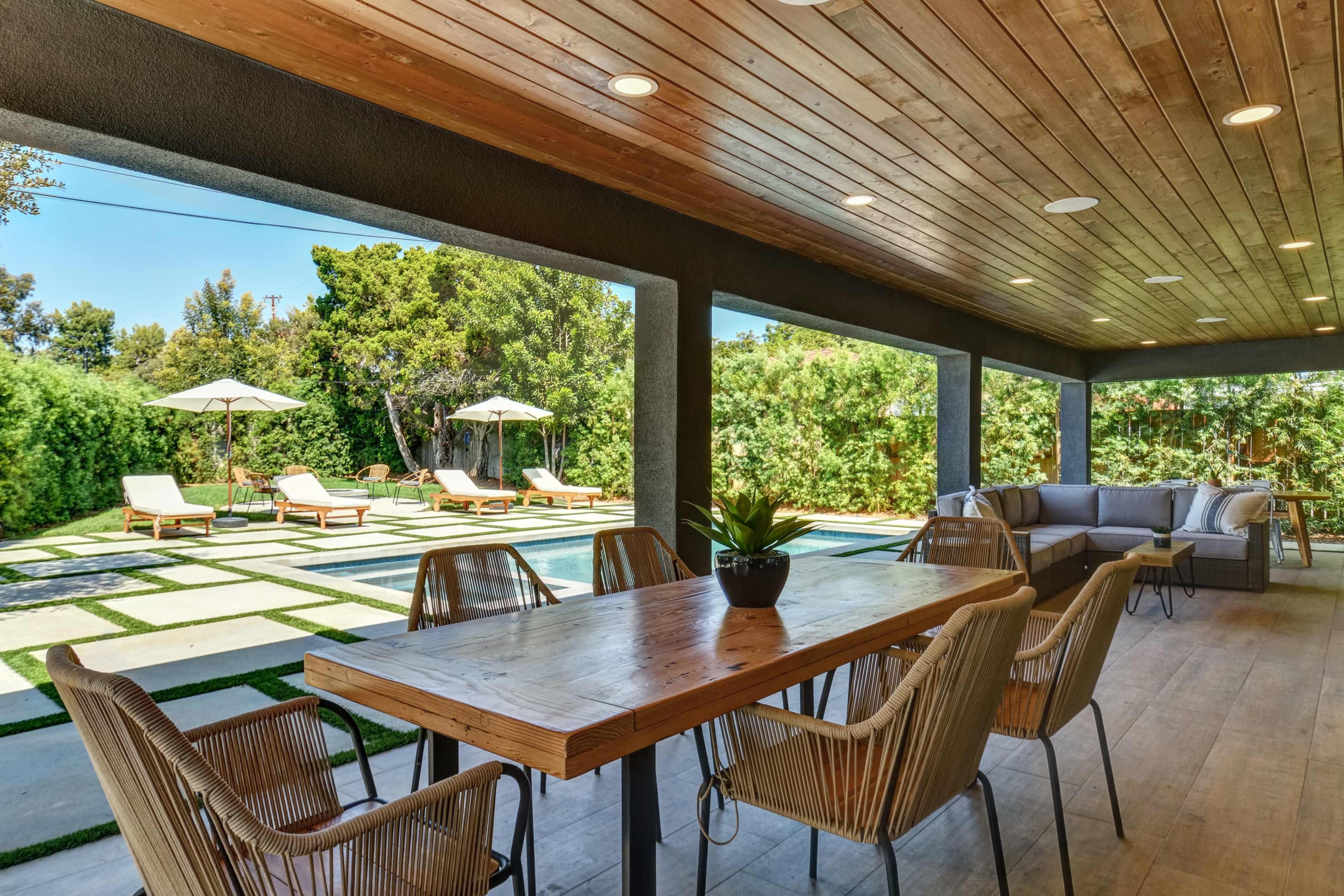 A covered patio features a large wooden dining table surrounded by chairs, with a view of a landscaped yard containing lounge chairs and umbrellas.