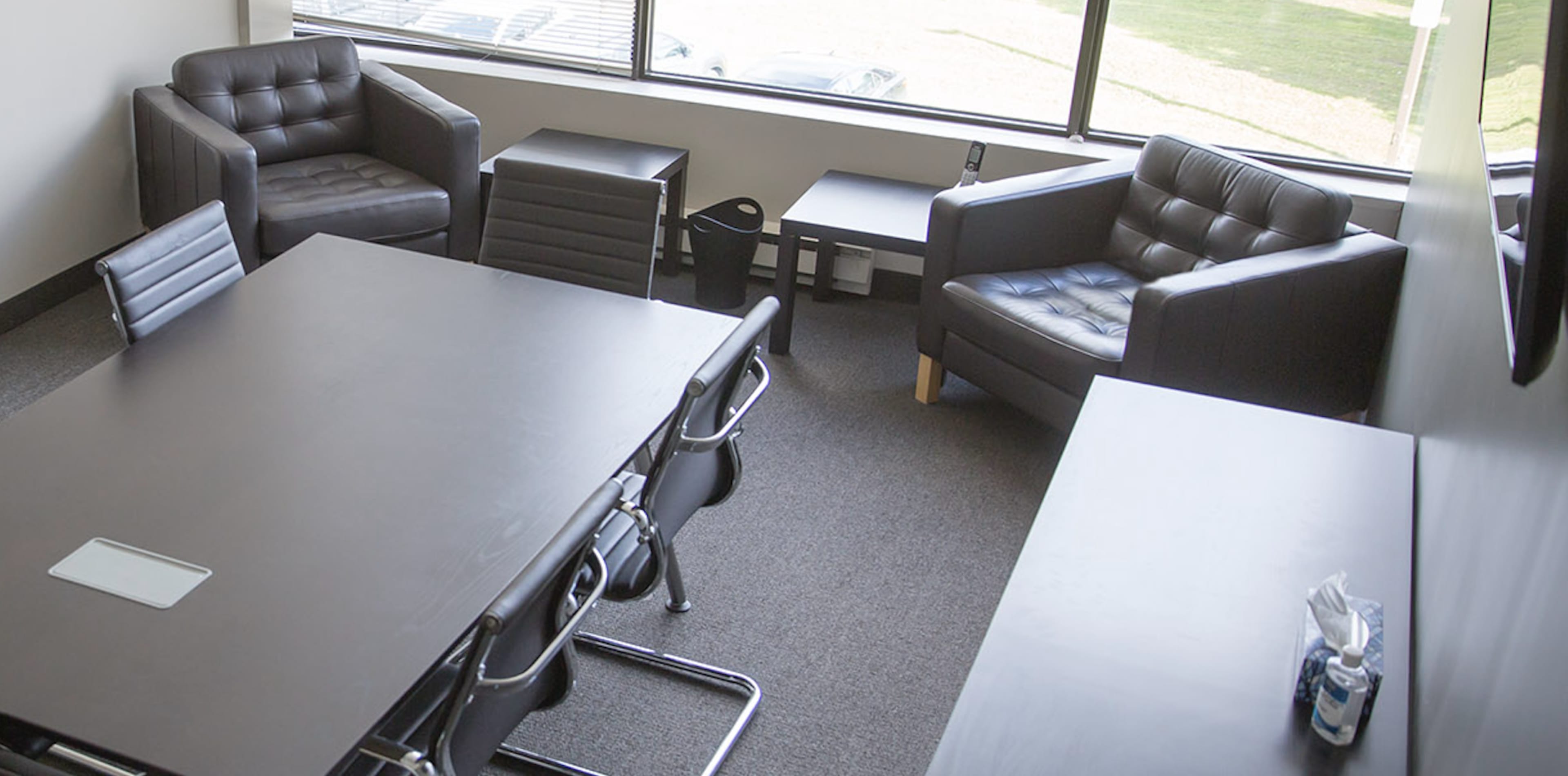 A modern conference room with a large black table, several metal chairs, and two leather armchairs beside a window.
