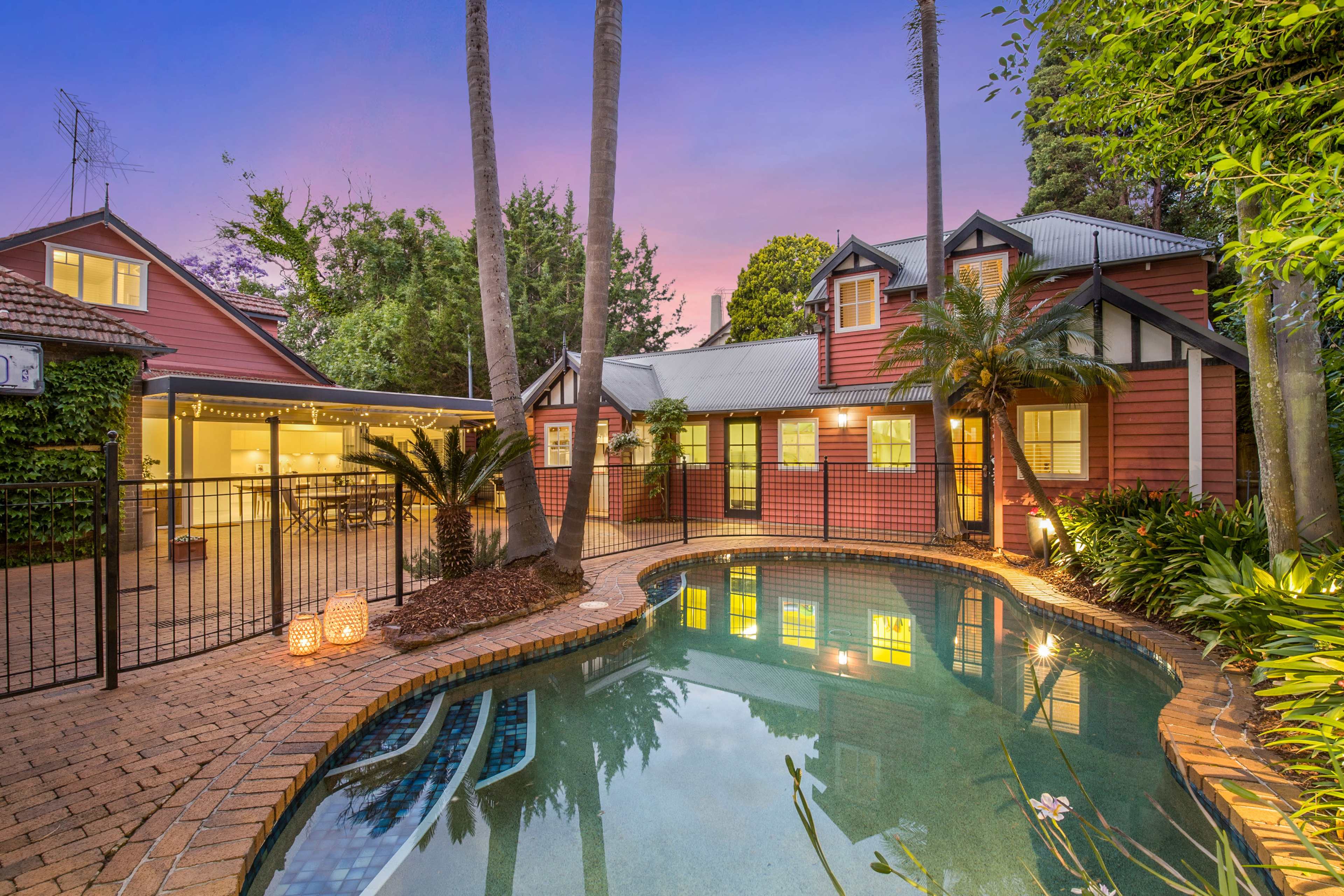 A backyard featuring a swimming pool surrounded by a red and white house, palm trees, and a dining area with outdoor seating at dusk.