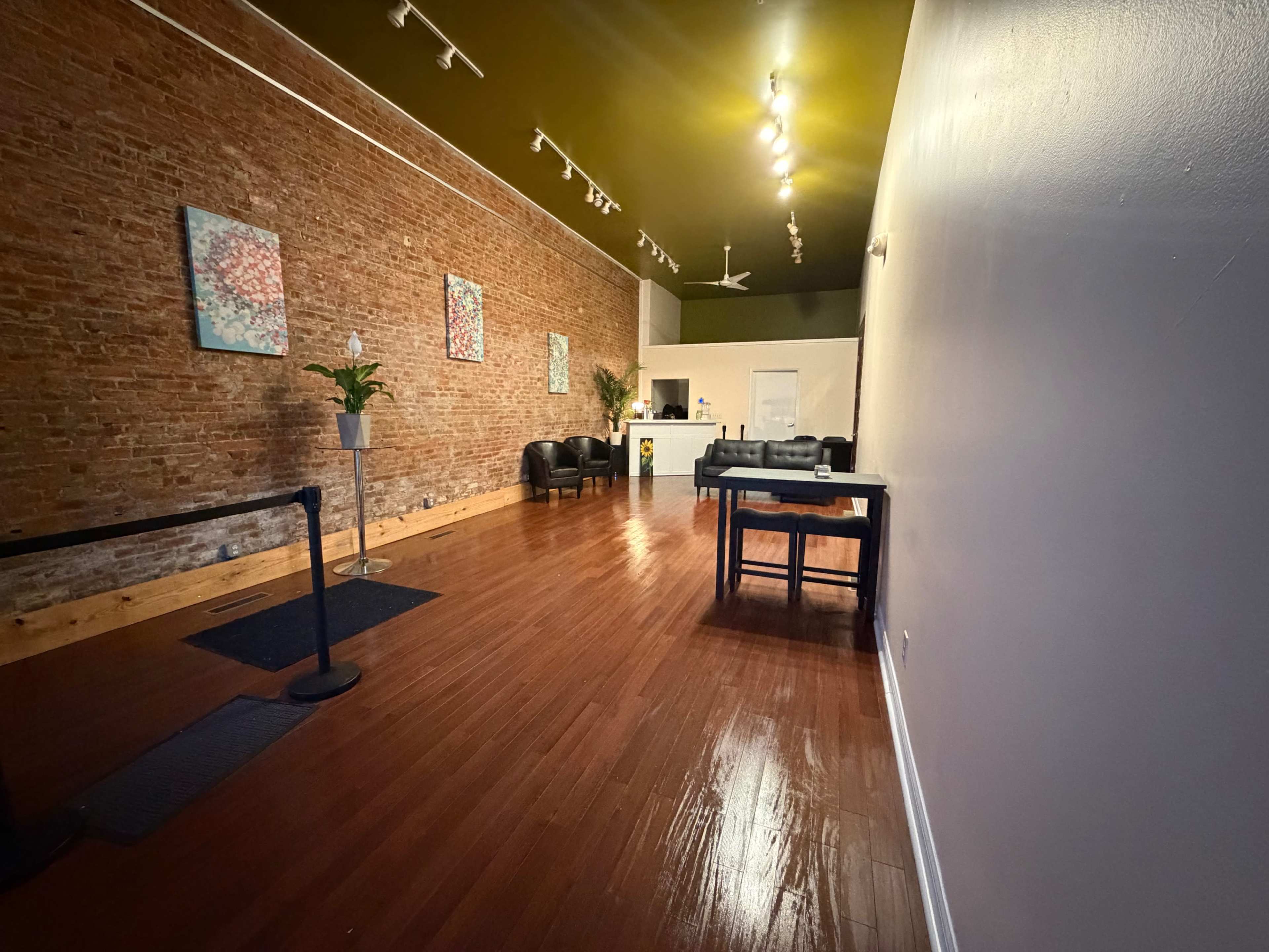 The image shows an empty, spacious interior with exposed brick walls, wooden flooring, a reception area at the back, and seating arrangements on the left side.
