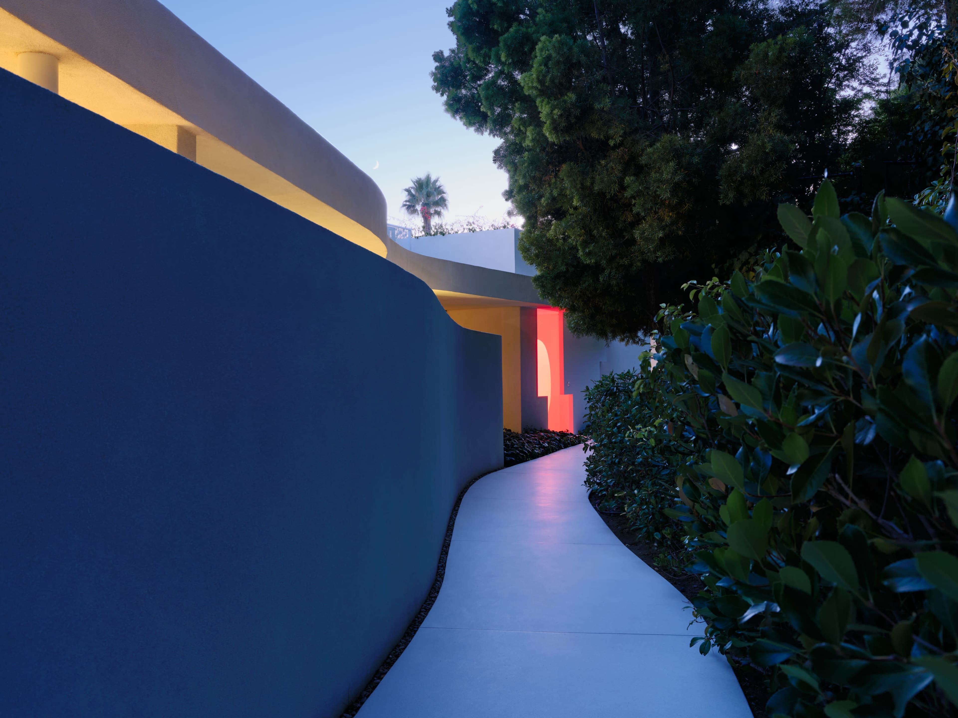 A curved pathway leads through greenery alongside a low wall illuminated by soft lights against a twilight sky.