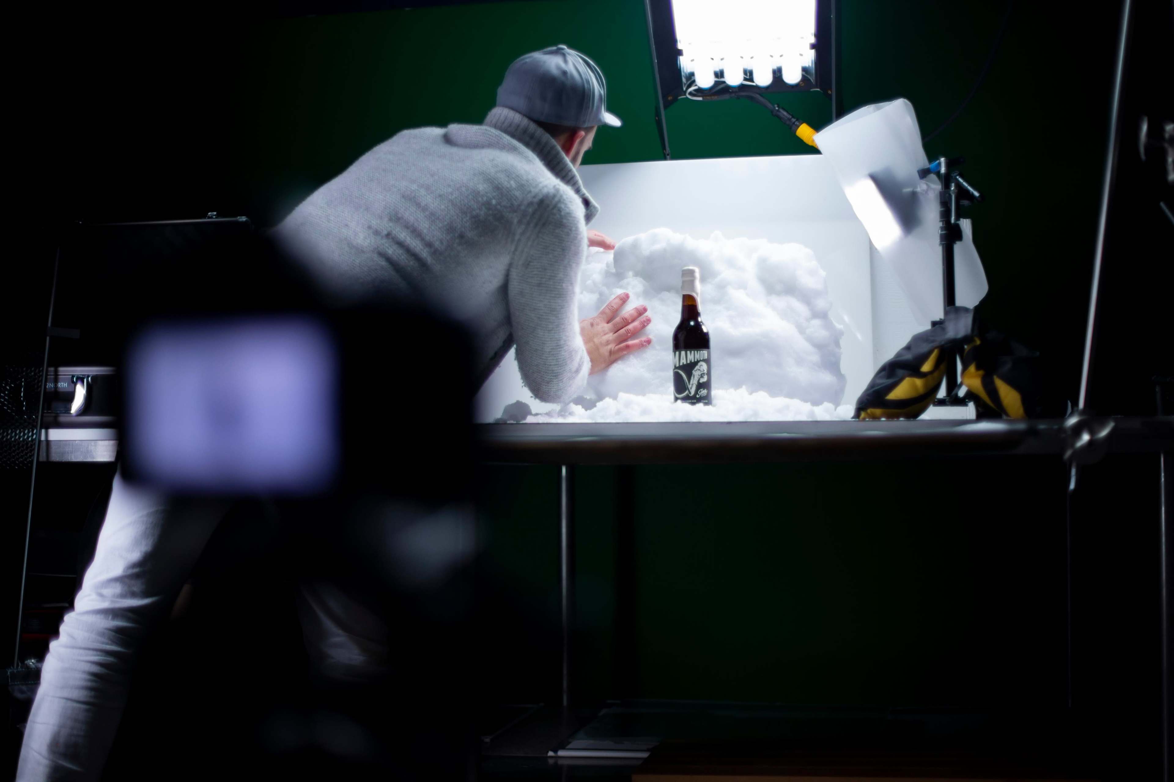 A person in a gray sweater and cap is arranging a snowy backdrop for a photography shoot featuring a bottle of beer on a table.