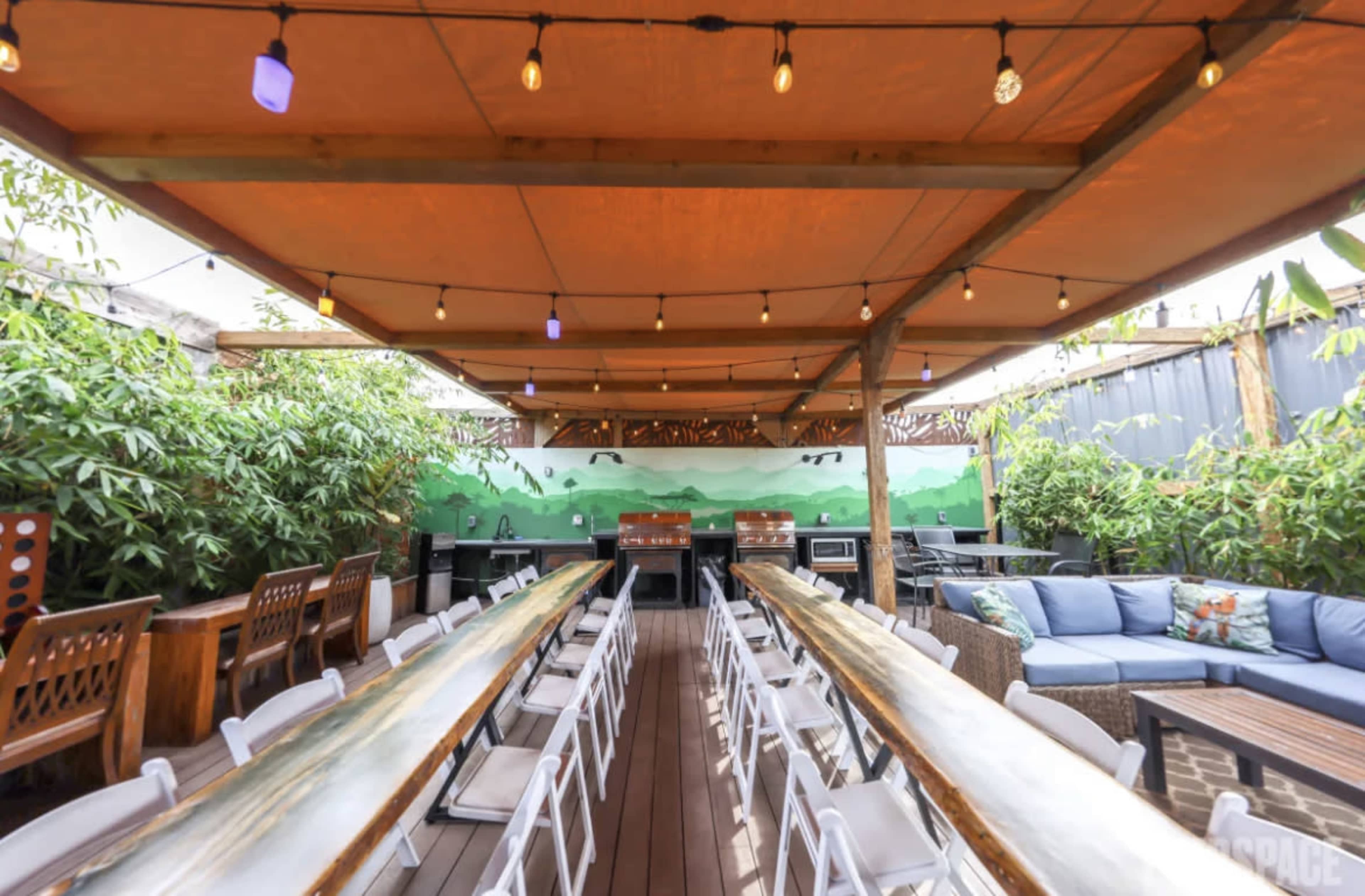 The image shows a rooftop dining area featuring long tables, string lights, and greenery along the walls.