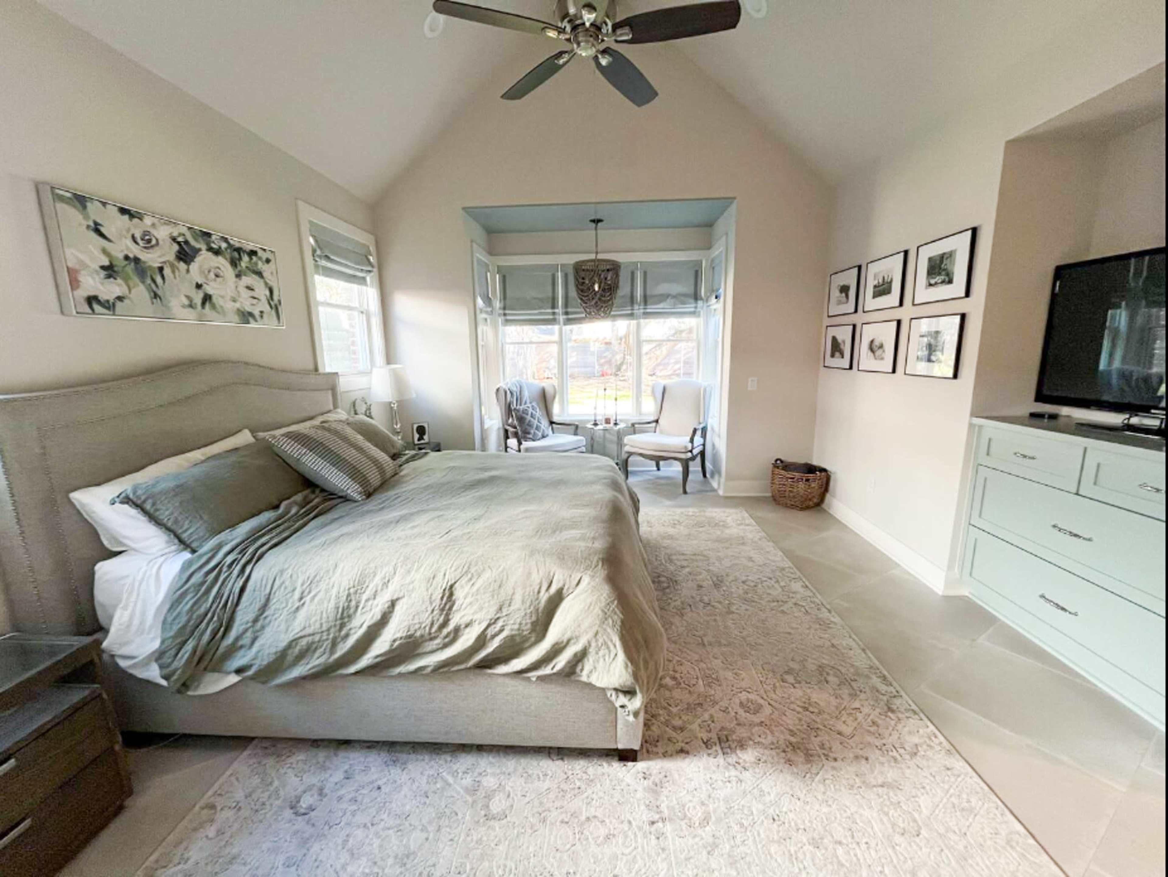A light-colored bedroom featuring a large bed, a sitting area with two chairs, and a structured dresser against the wall.