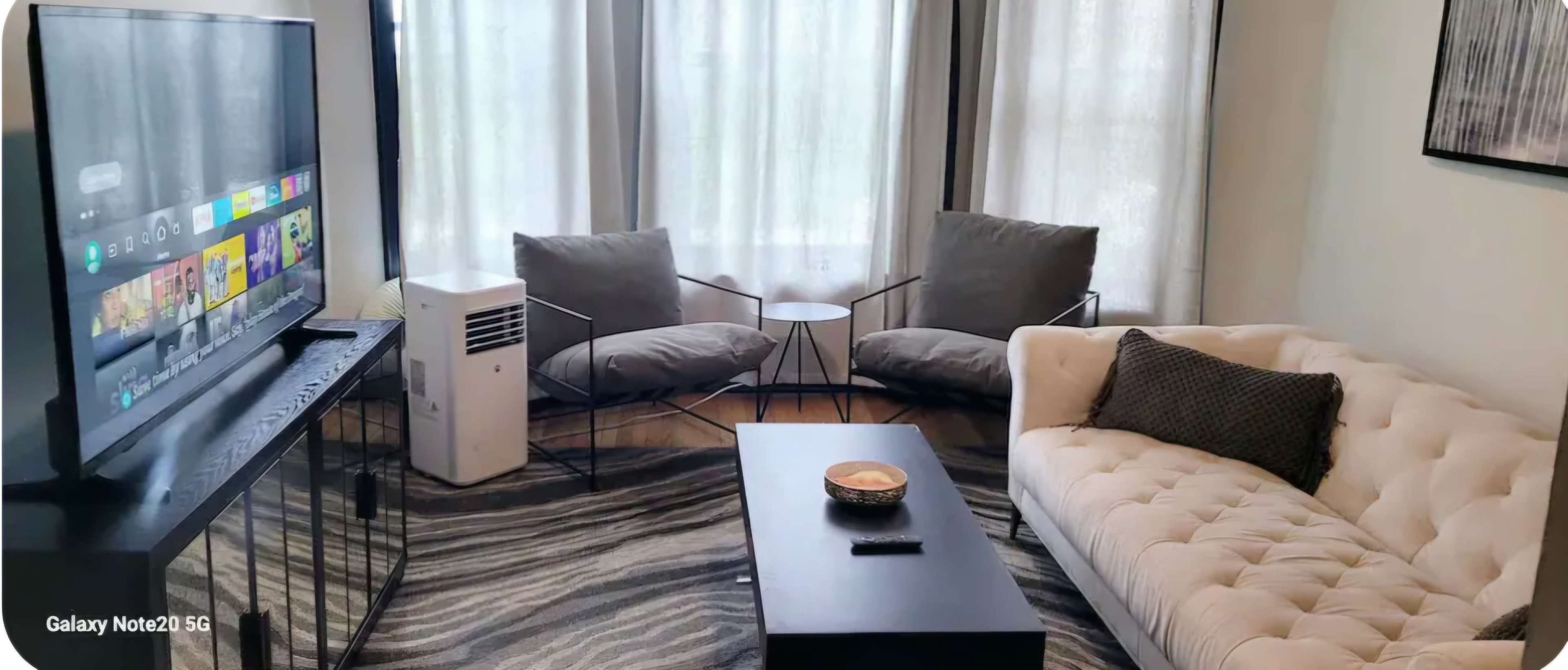 The image shows a living room with a beige sofa, two gray armchairs, a small side table, and a television on a stand, all set on a patterned rug.