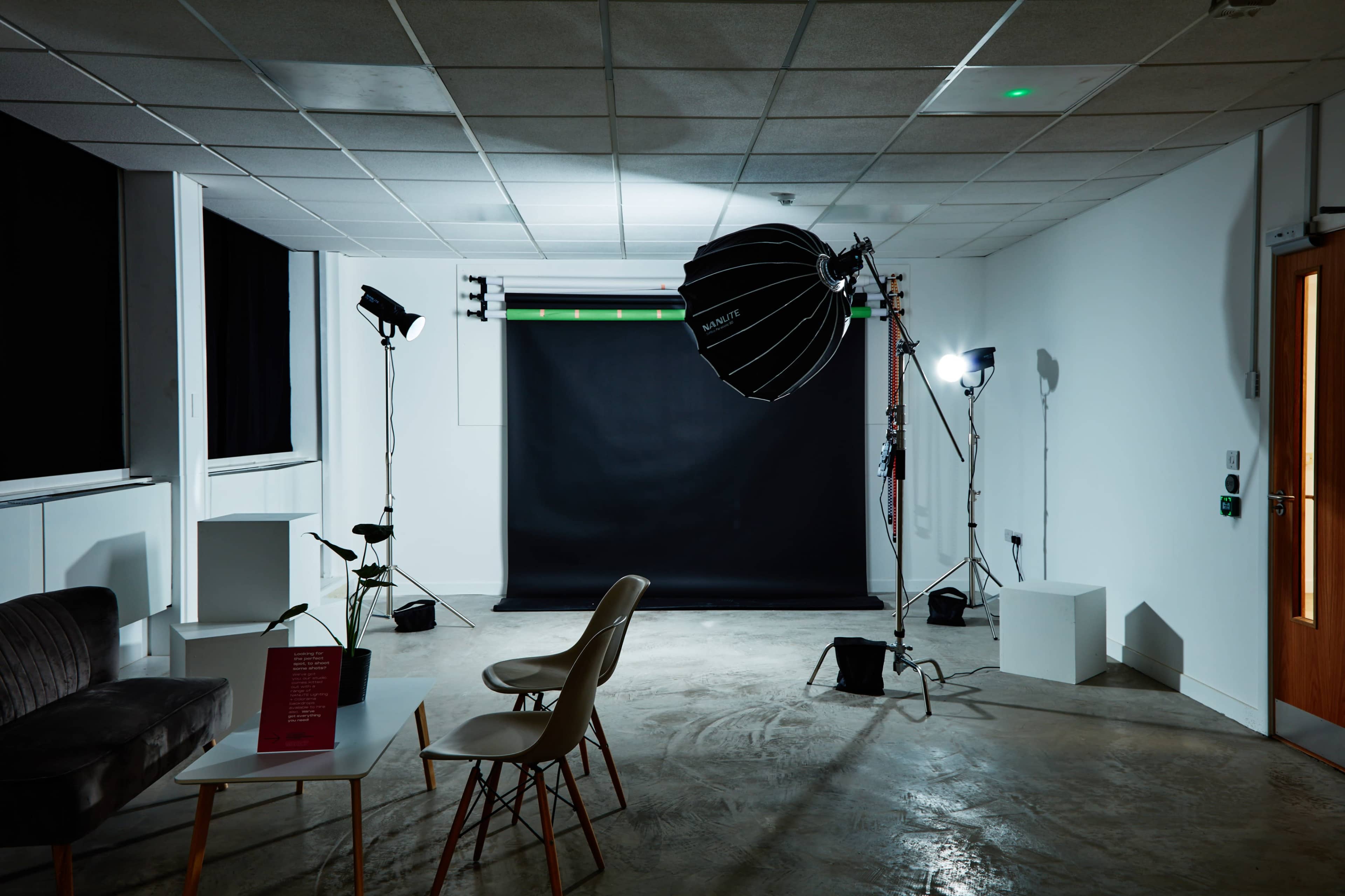 The image shows a photography studio with a black backdrop, various lighting equipment, and a few pieces of furniture including chairs and a table.