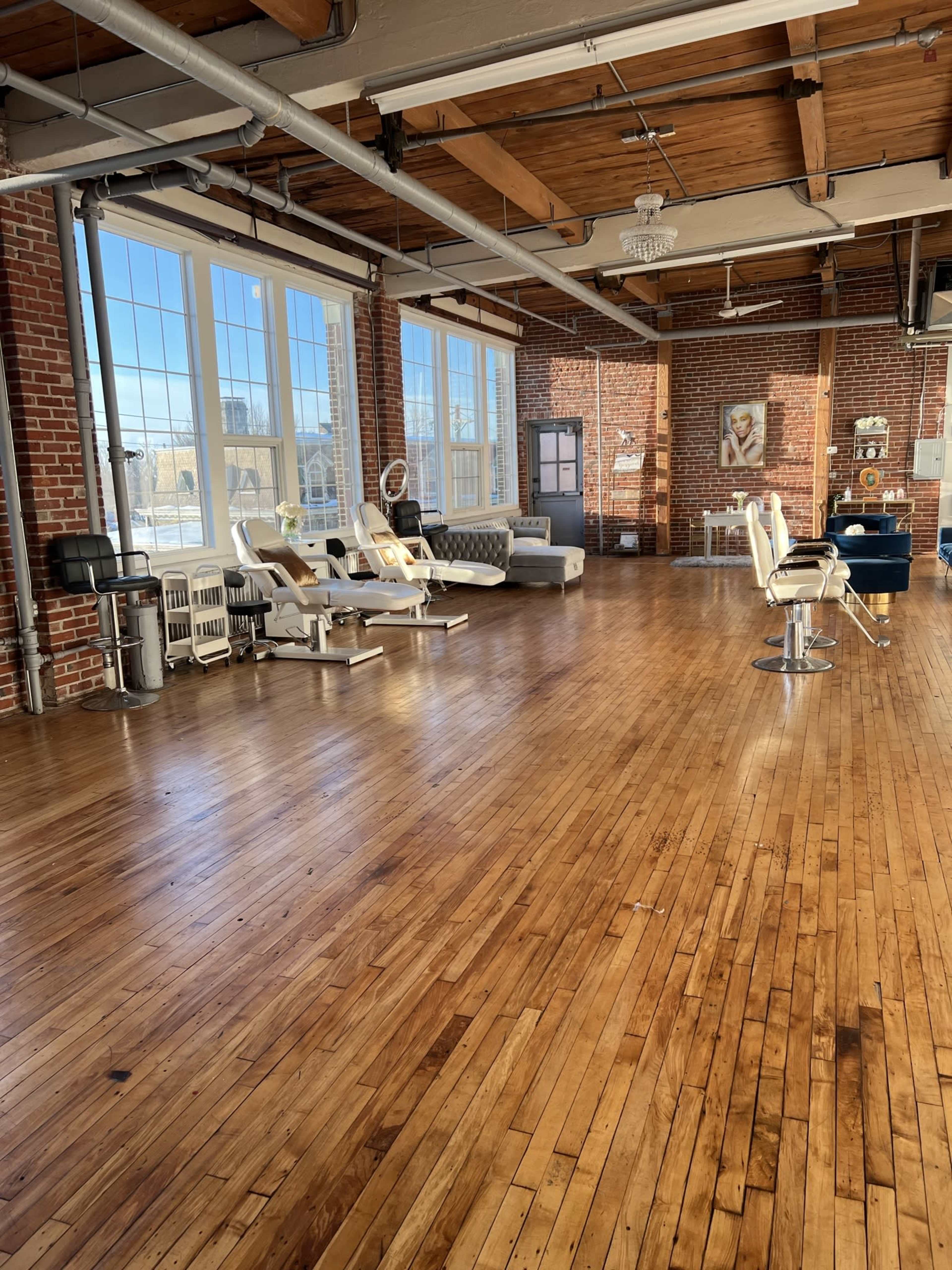 The image shows a spacious, well-lit interior with hardwood floors, featuring salon chairs, treatment beds, and large windows with a view.