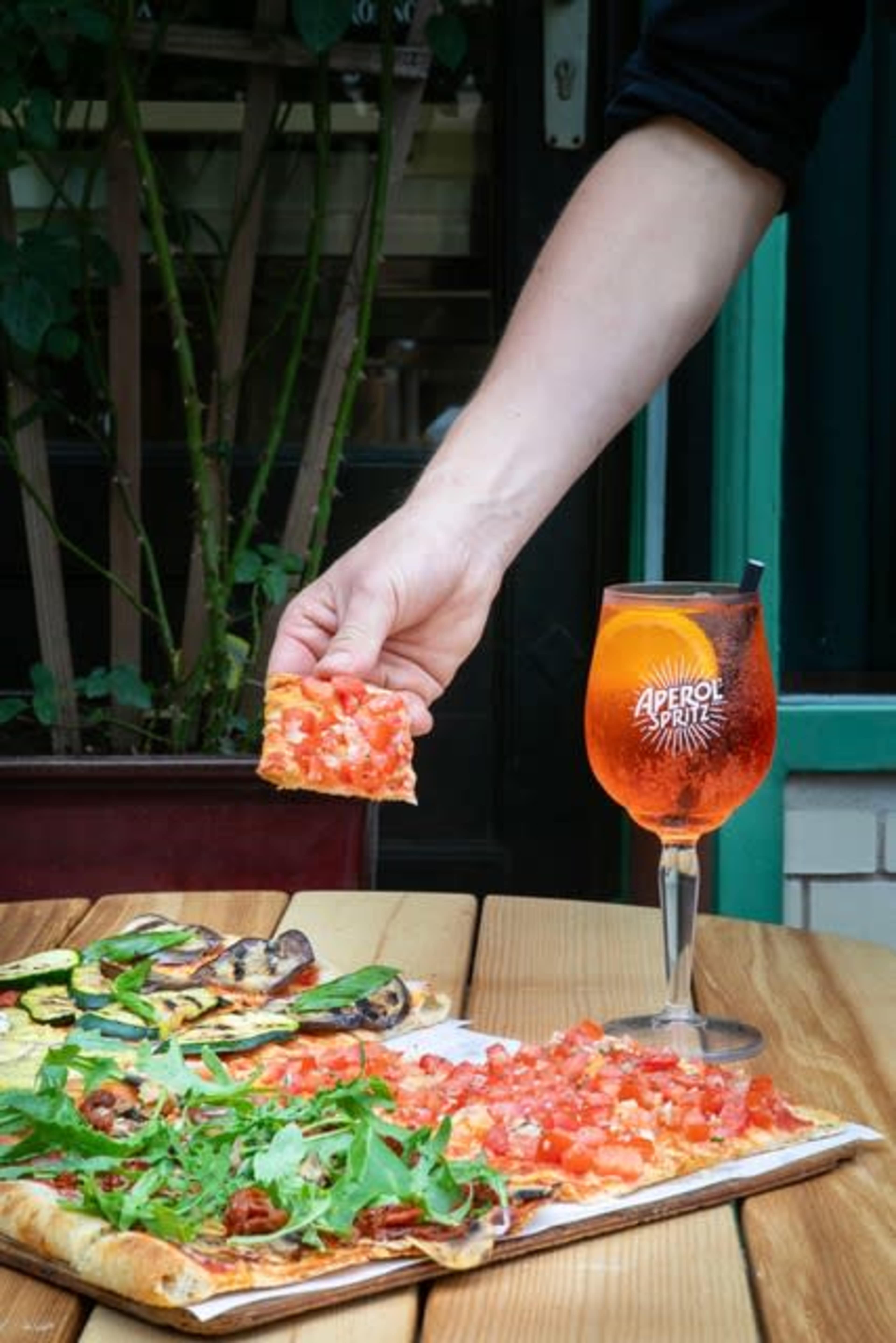 A person holds a slice of pizza topped with tomatoes over a wooden table, next to a glass of Aperol Spritz.