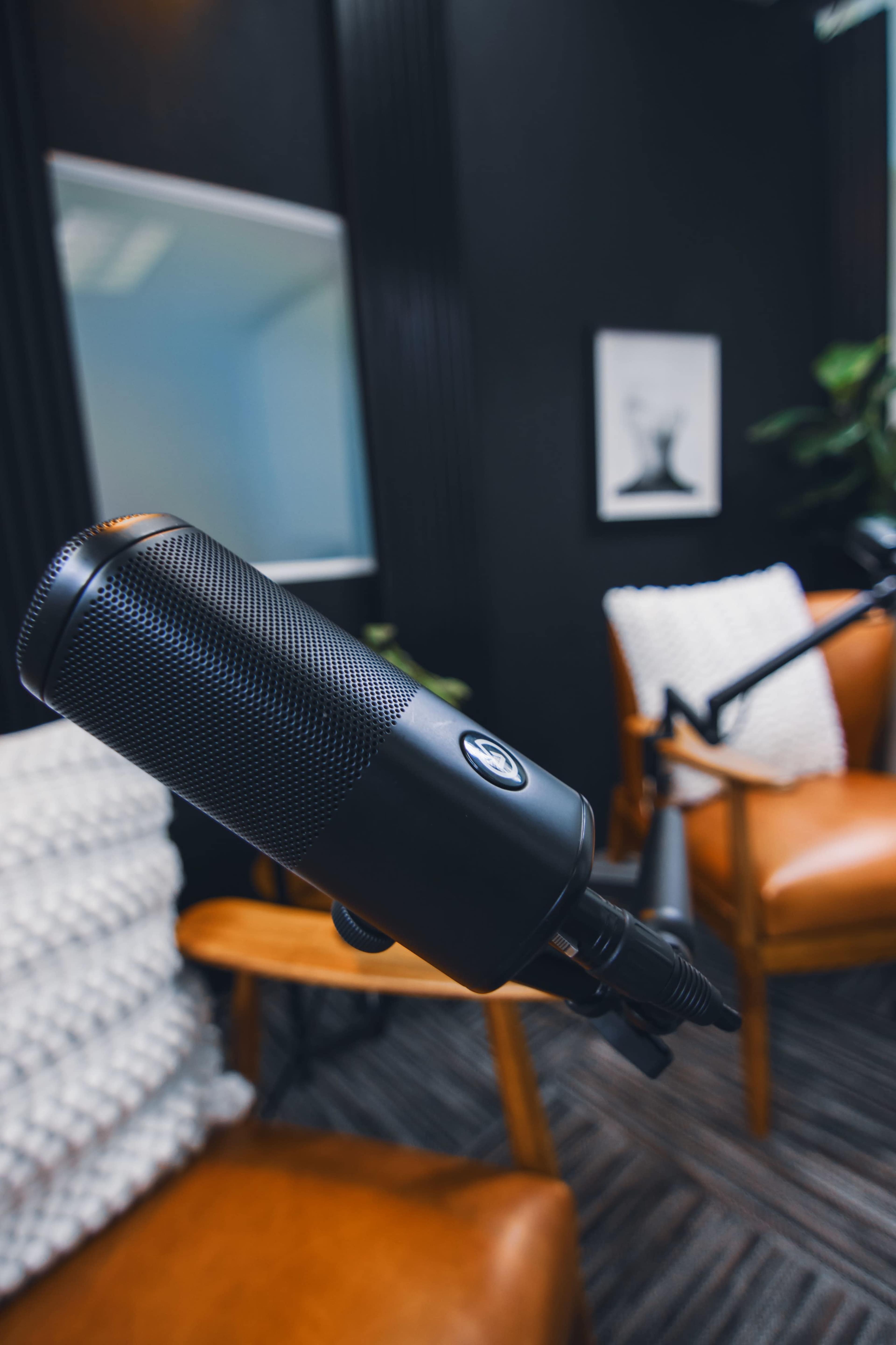 A close-up of a black microphone positioned in a modern room with wooden chairs and a soft, textured backdrop.
