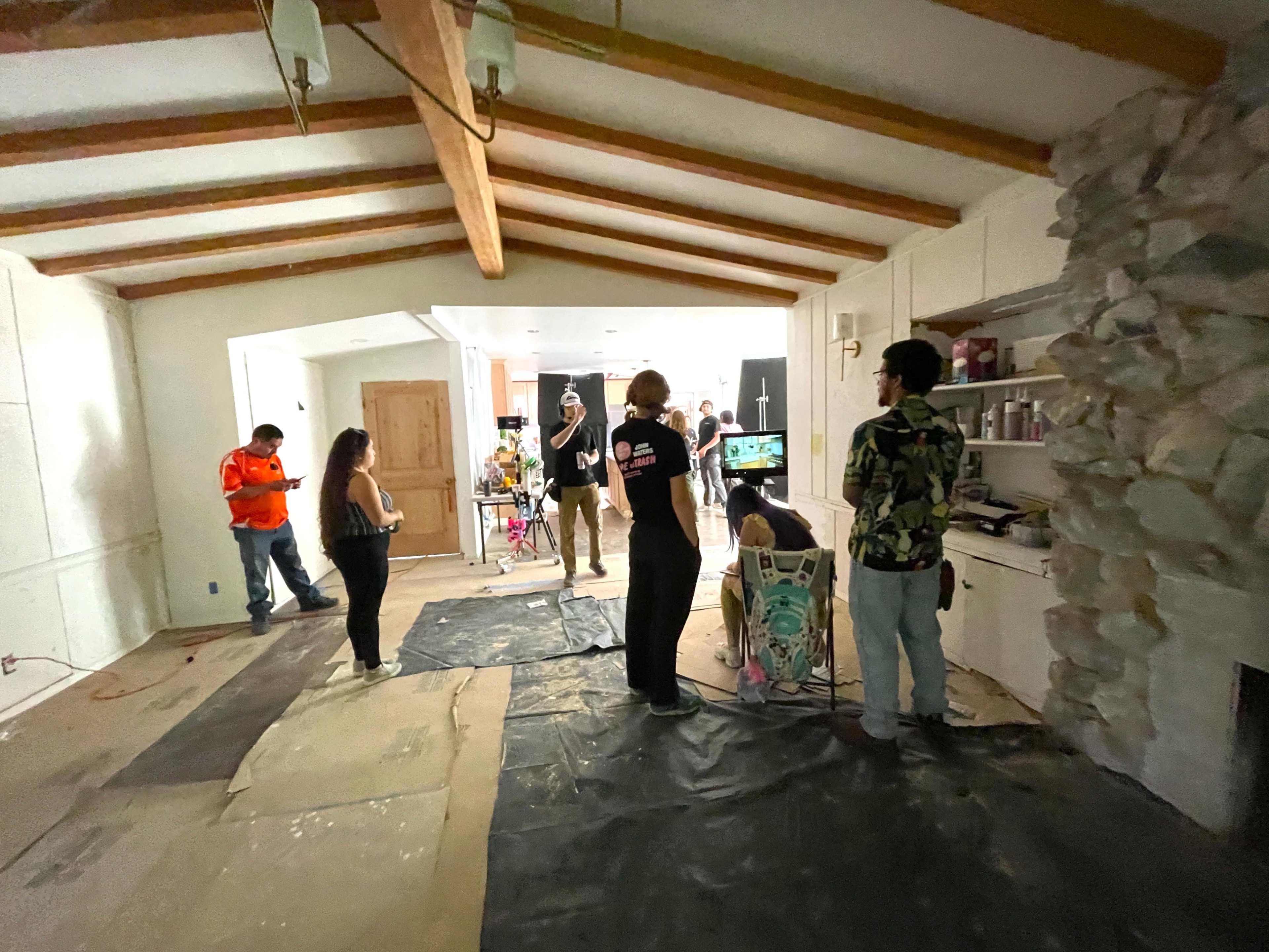 A group of people is gathered in a partially renovated room with wooden beams and a stone wall, while working on various tasks and using equipment.