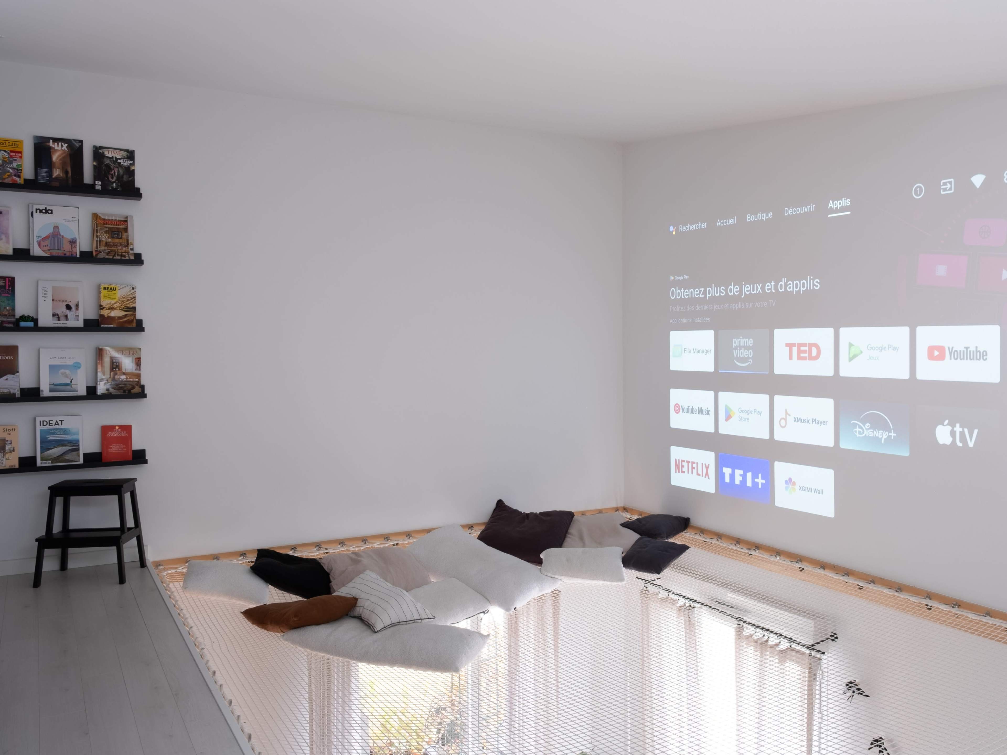 A minimalist room features a projector screen displaying various streaming service logos, with a netted seating area filled with cushions and a shelf of books on one wall.