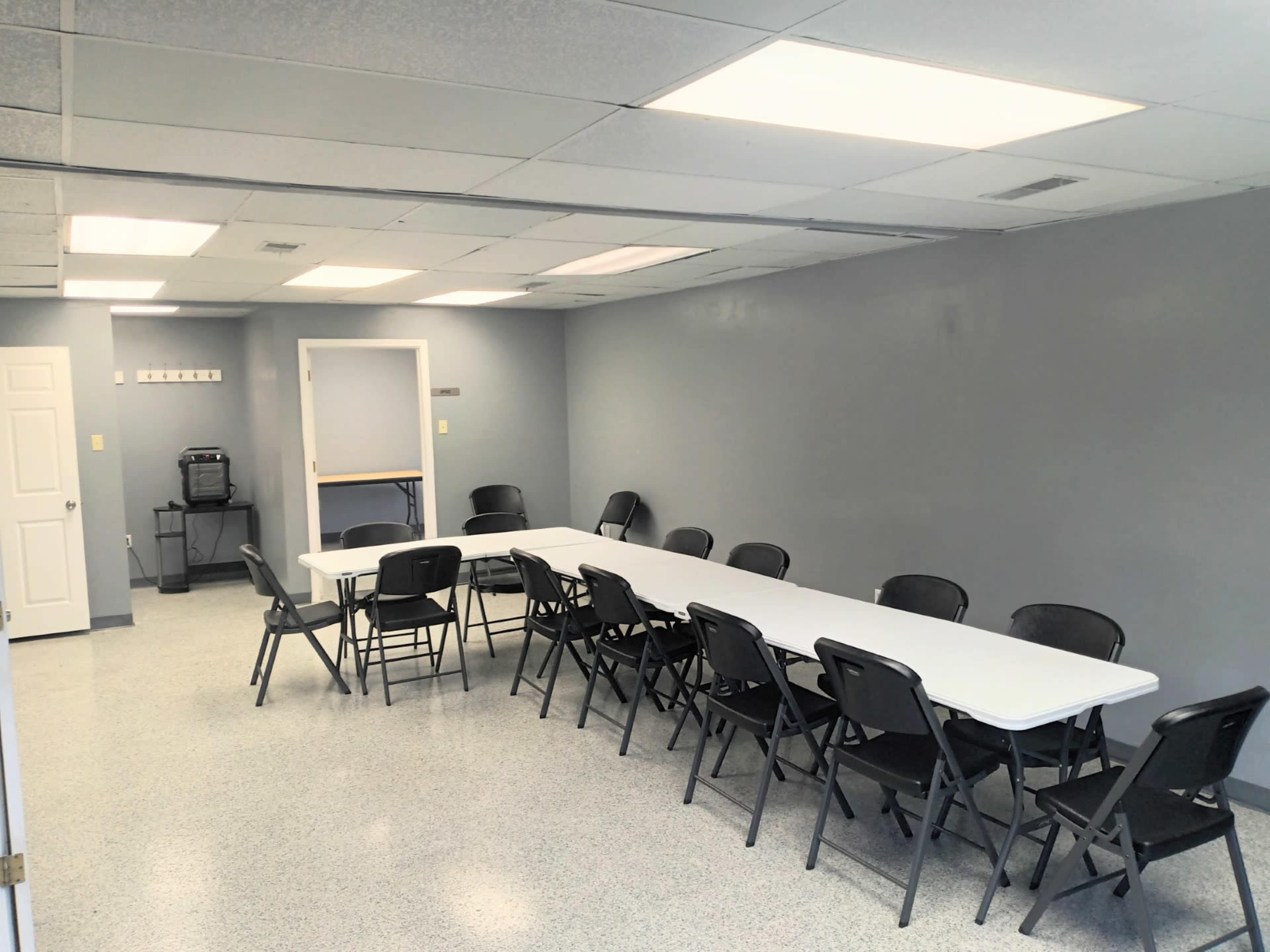 A large, empty meeting room features a long white table surrounded by black chairs, with a kitchenette area in the background.