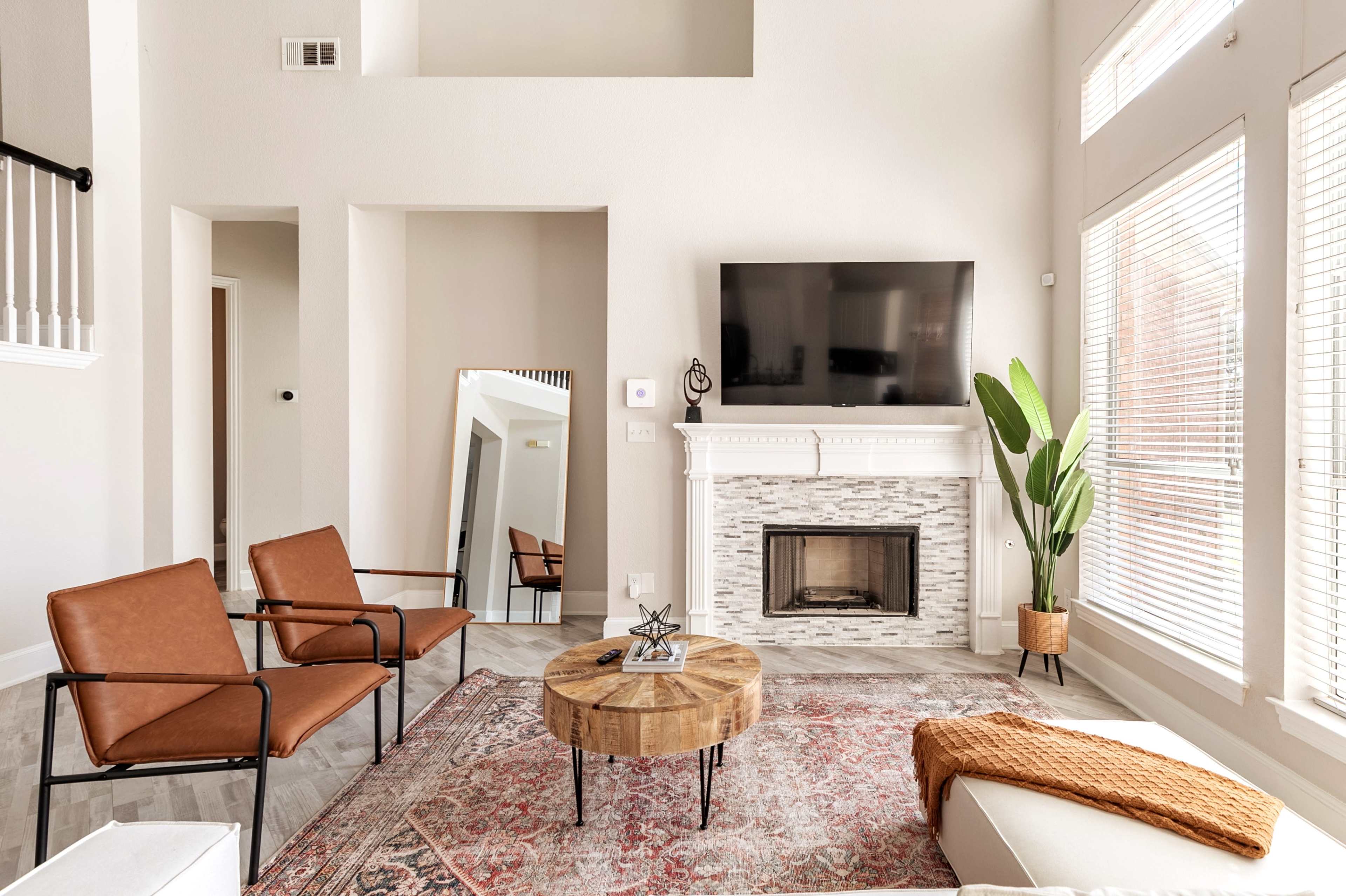 A living room features two brown chairs, a round wooden coffee table, a fireplace, and a large television mounted on a light-colored wall with large windows.