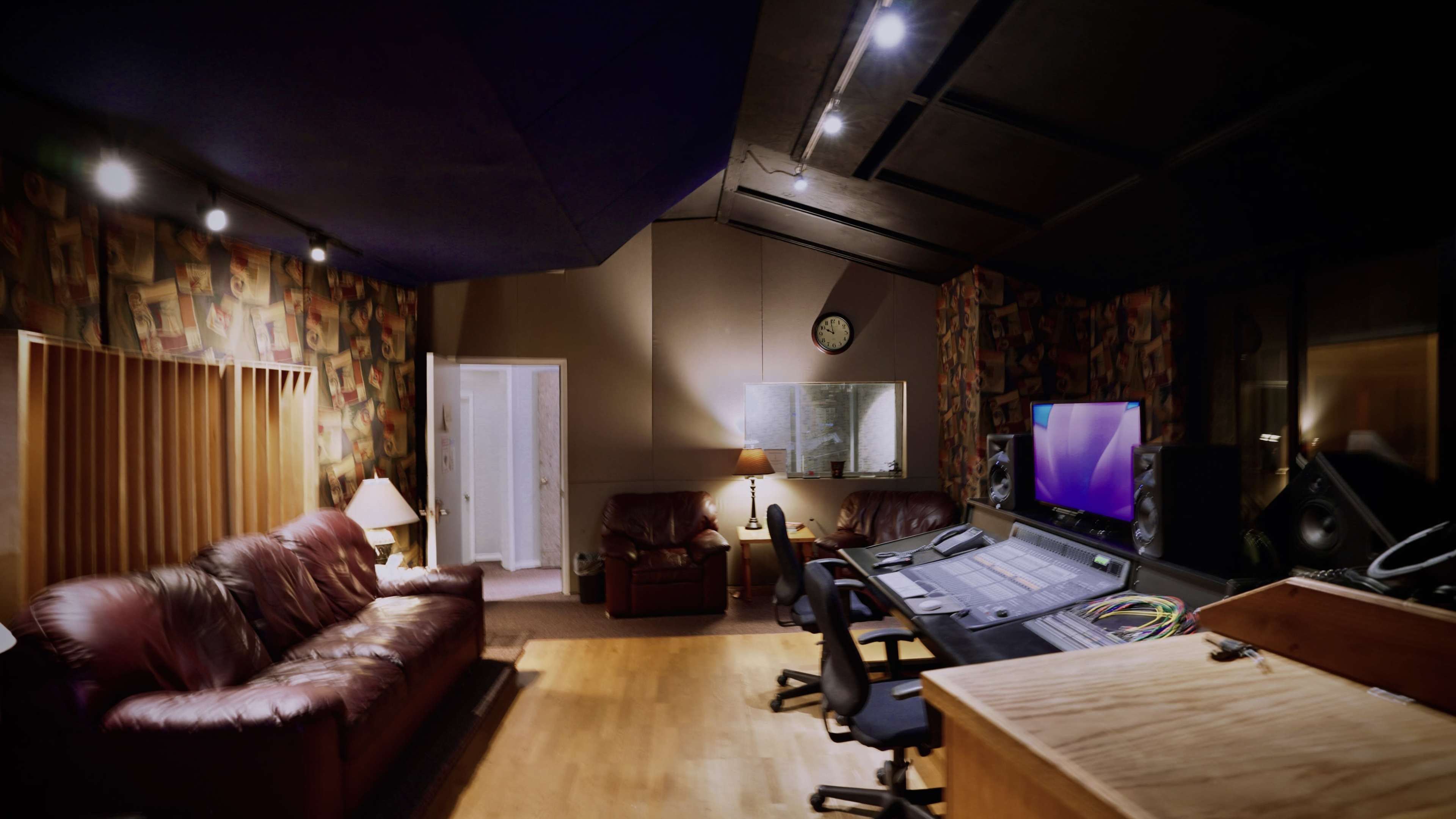 A sound recording studio features a control area with a mixing console, two chairs, and a couch, complemented by patterned walls and soft lighting.