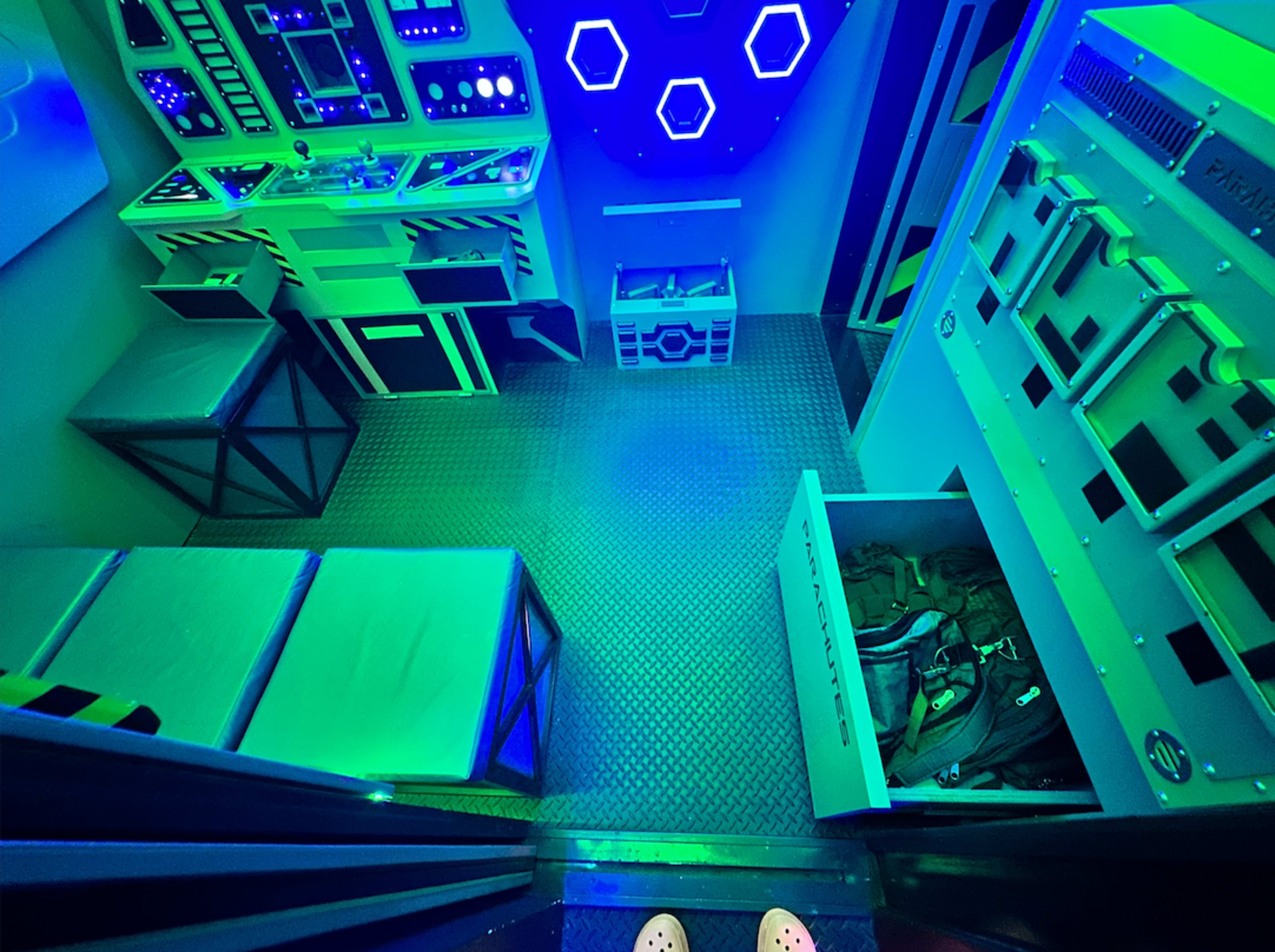 The image shows a futuristic room with green and blue lighting, featuring a control panel, storage crates, and two beds on the floor.