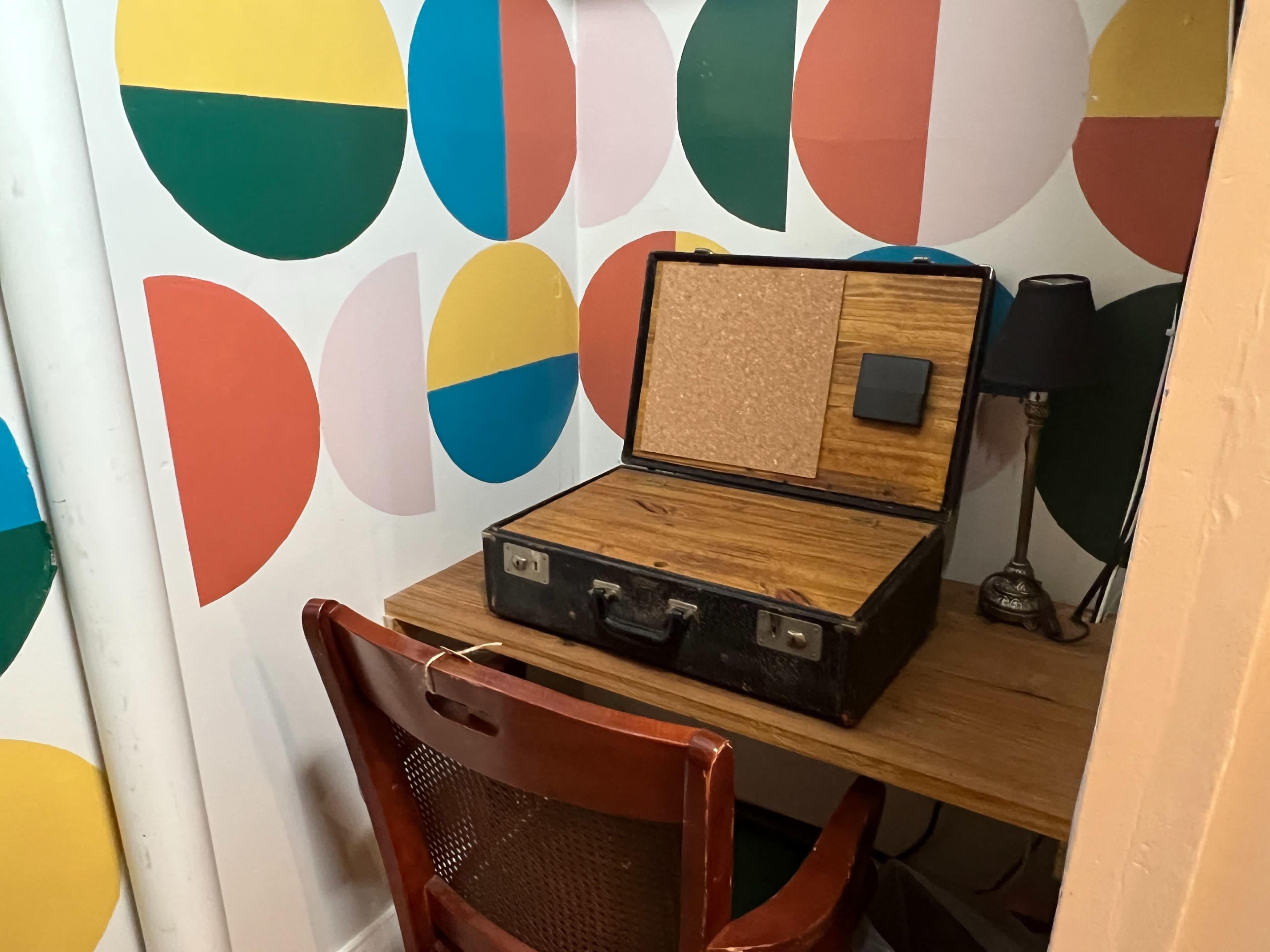 A wooden desk with a chair sits beside an open vintage suitcase, placed against a wall decorated with colorful geometric shapes.