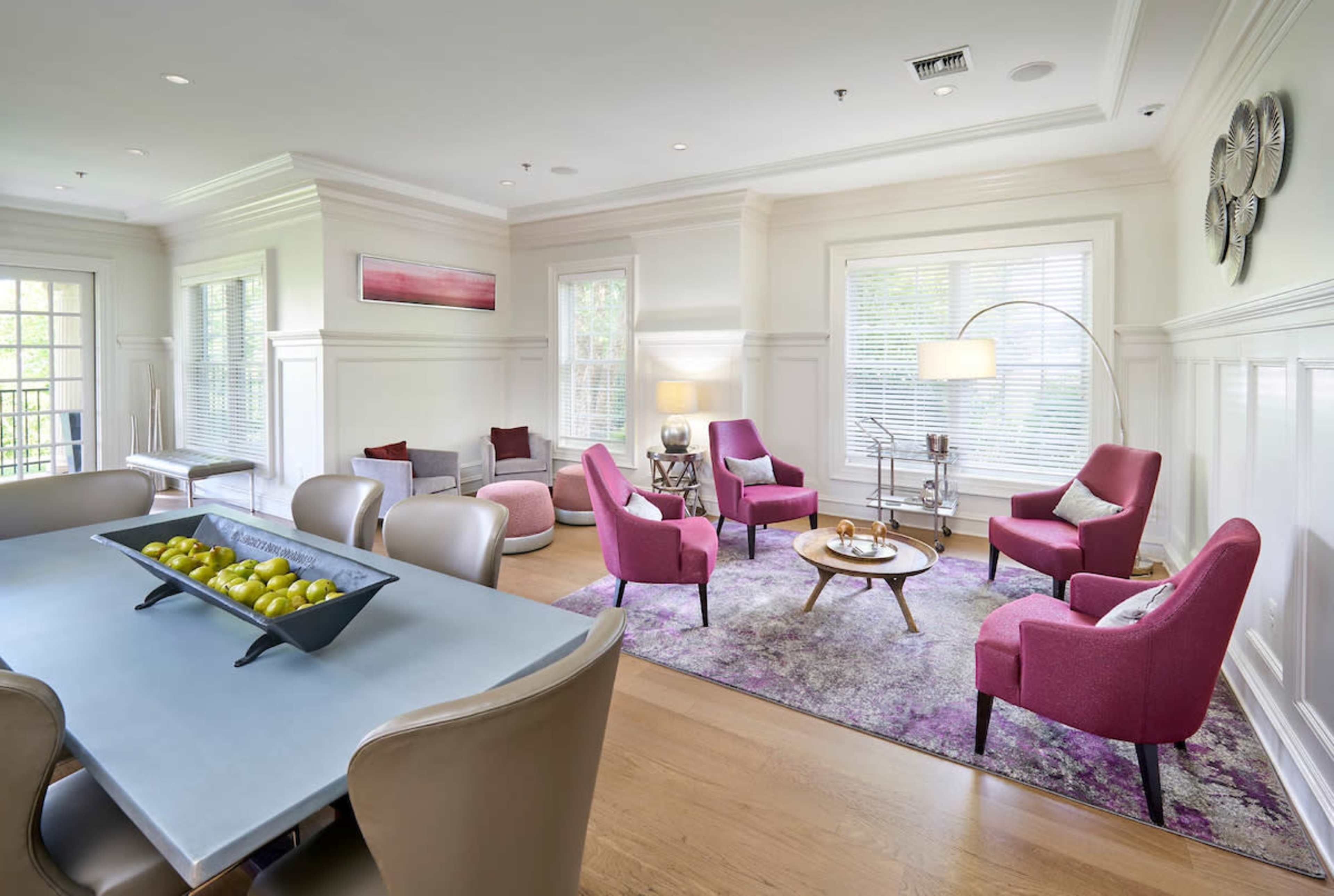 The room features a mix of modern seating arrangements with pink chairs, a coffee table, and a dining area, all set against light-colored walls and hardwood flooring.