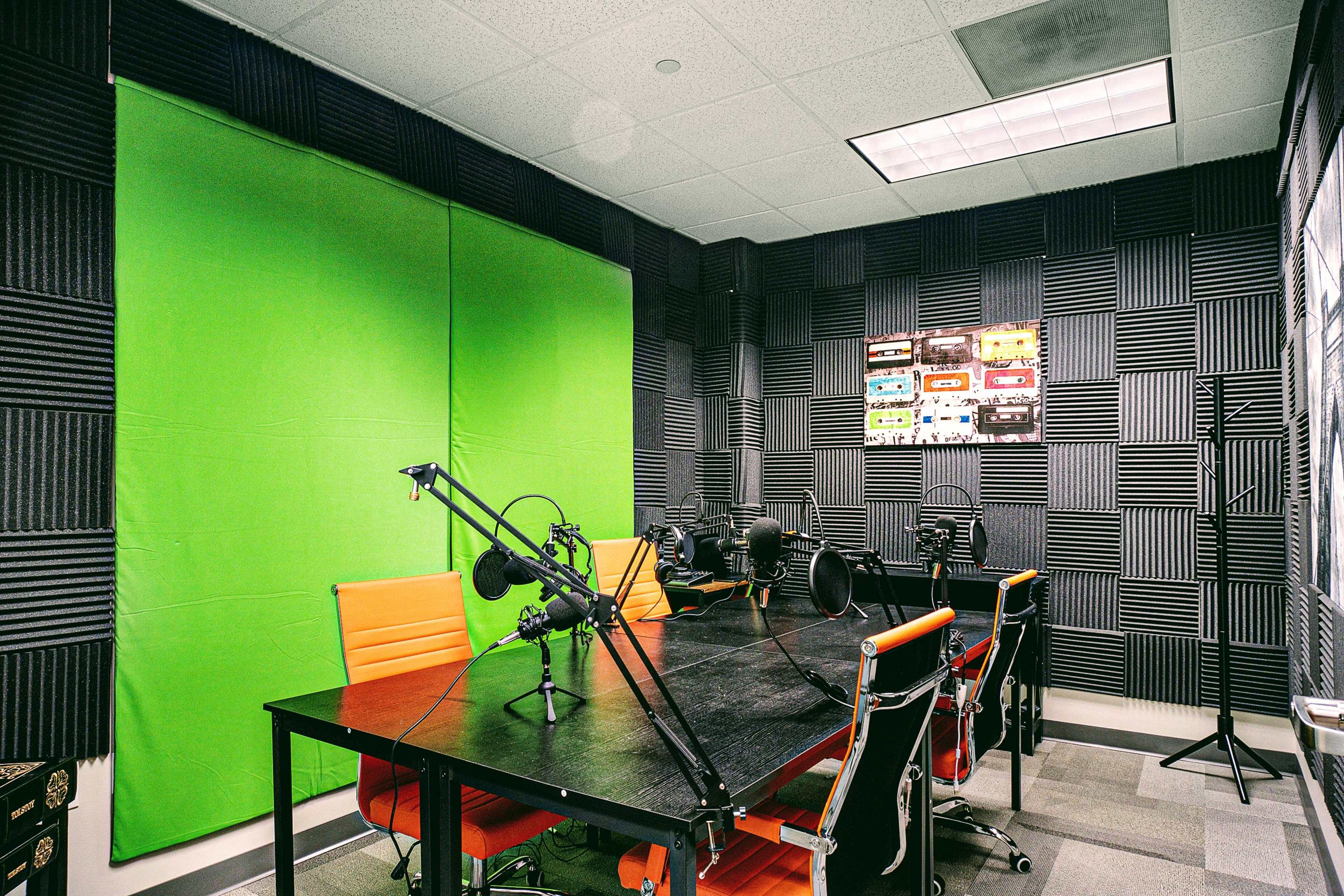A soundproof recording studio with a green backdrop, a large table, multiple microphones, and orange chairs.