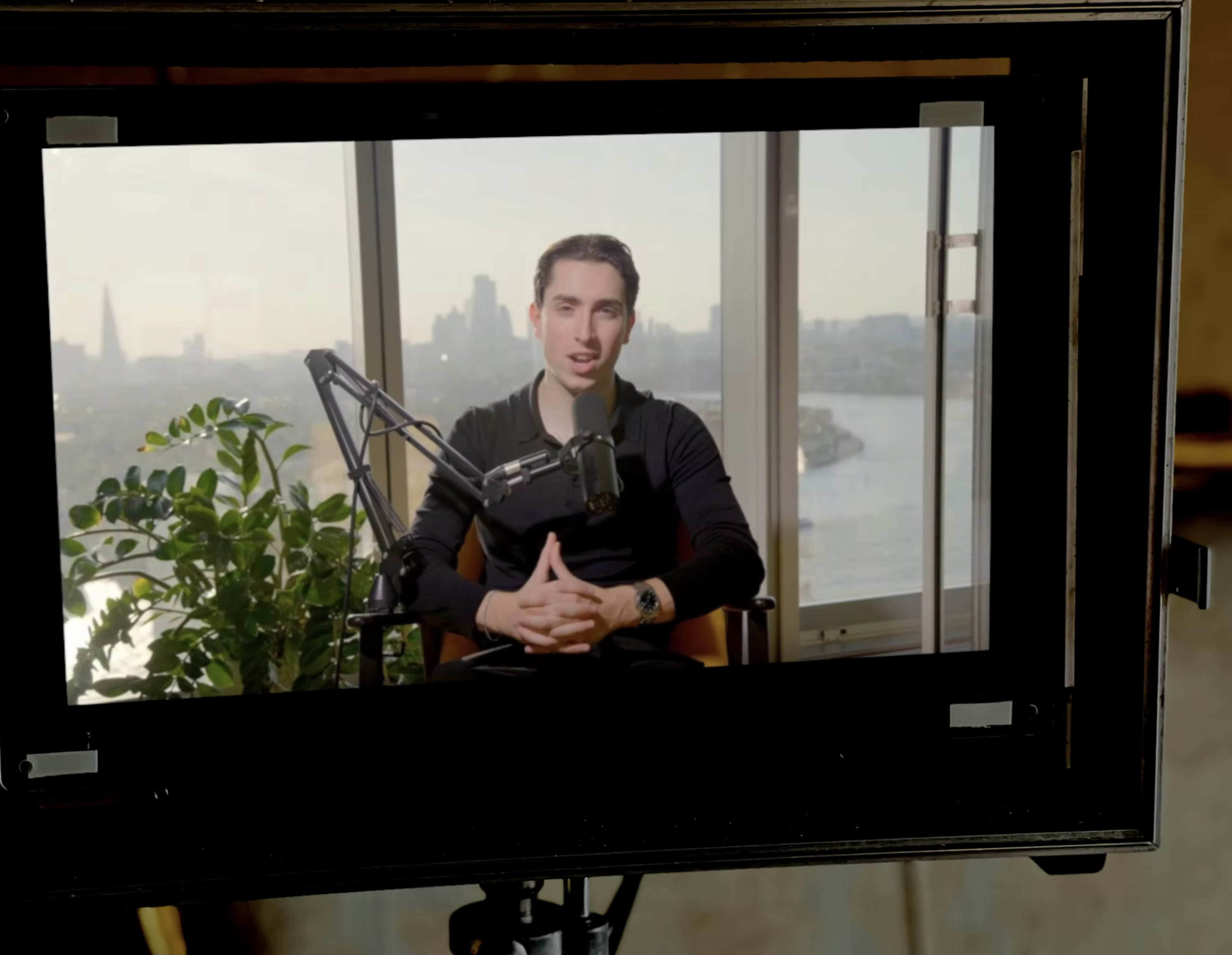 A man sits in front of a microphone and camera, speaking in a well-lit room with a city skyline visible through the large window behind him.