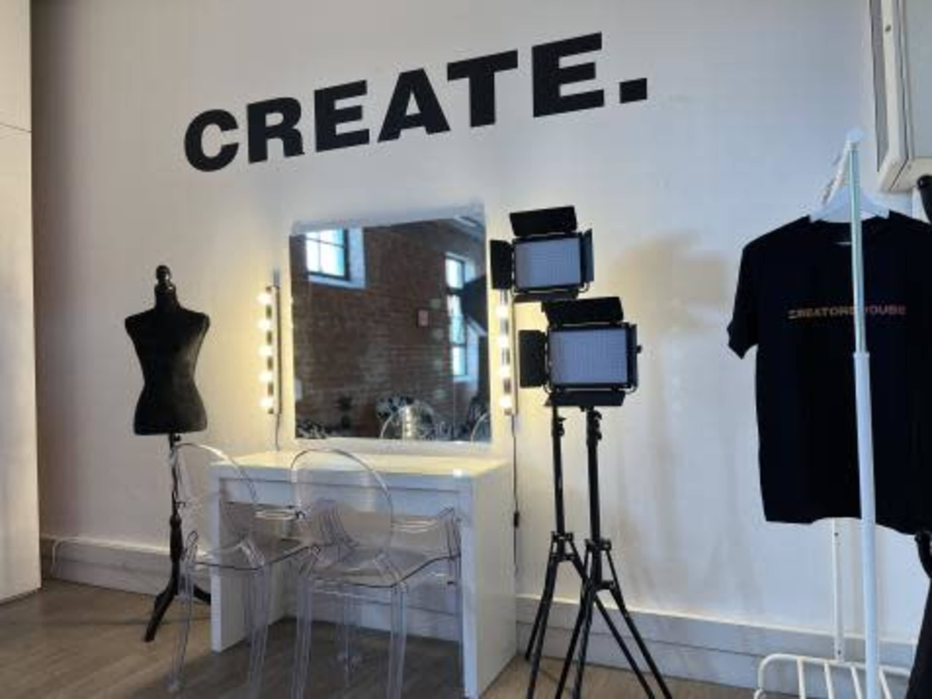 A well-lit vanity area with a mirror, two spotlight stands, a dress form, and a shirt on a rack against a wall that features the word "CREATE."