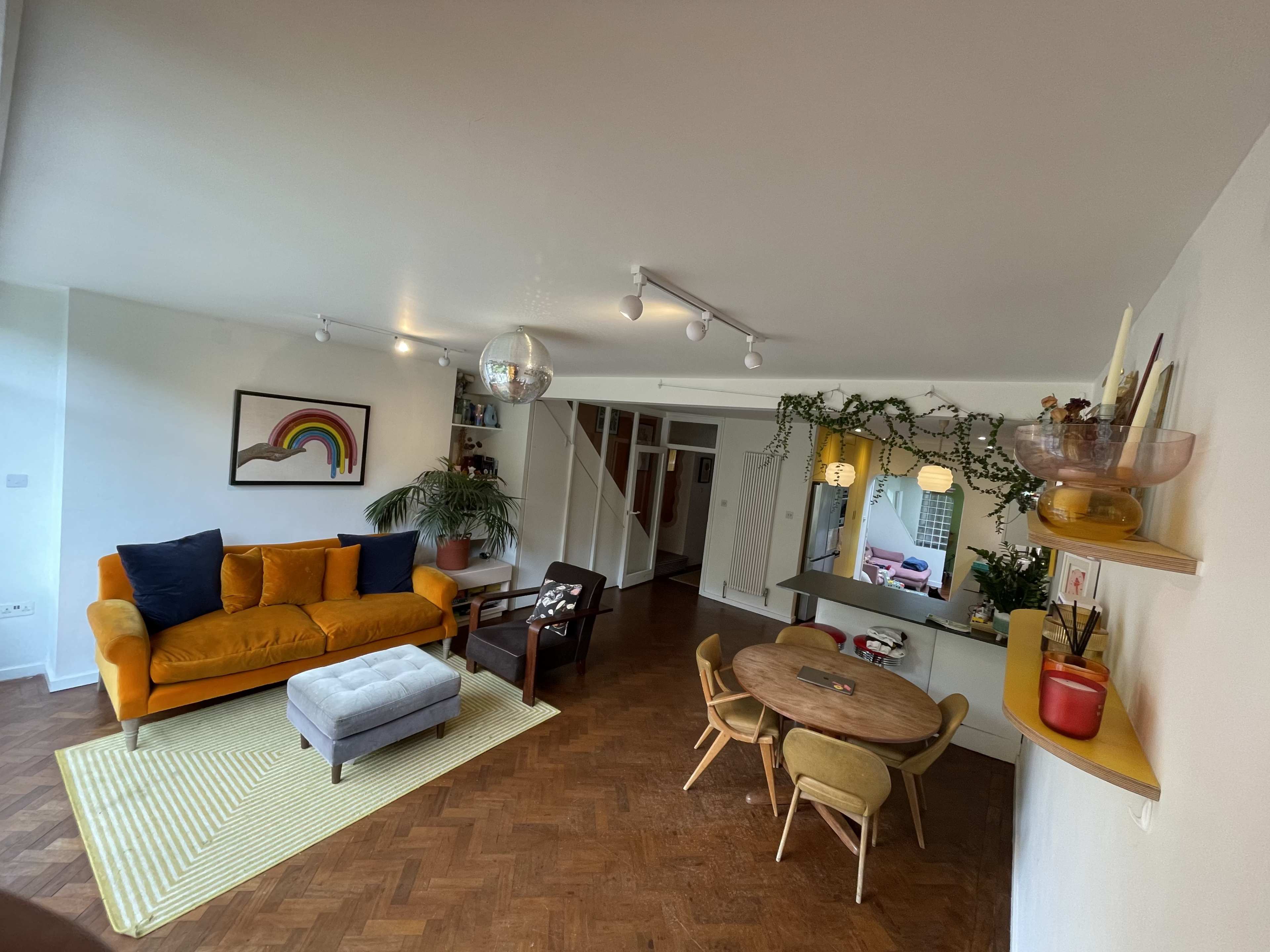 A cozy living room features a yellow sofa, a gray ottoman, a round dining table with chairs, and decor including a rainbow painting and indoor plants.