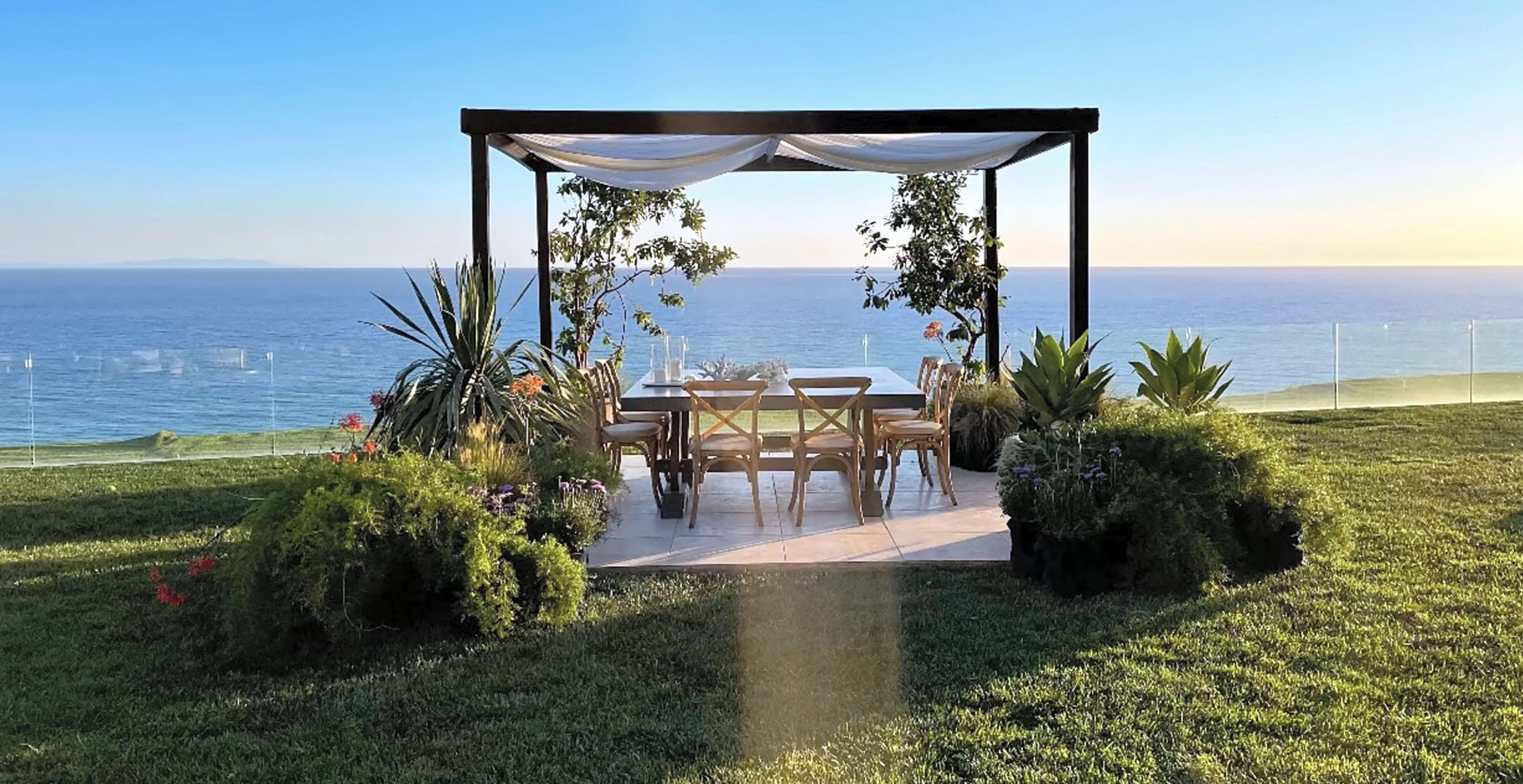 A wooden gazebo with a table and chairs sits on a grassy area overlooking the ocean.