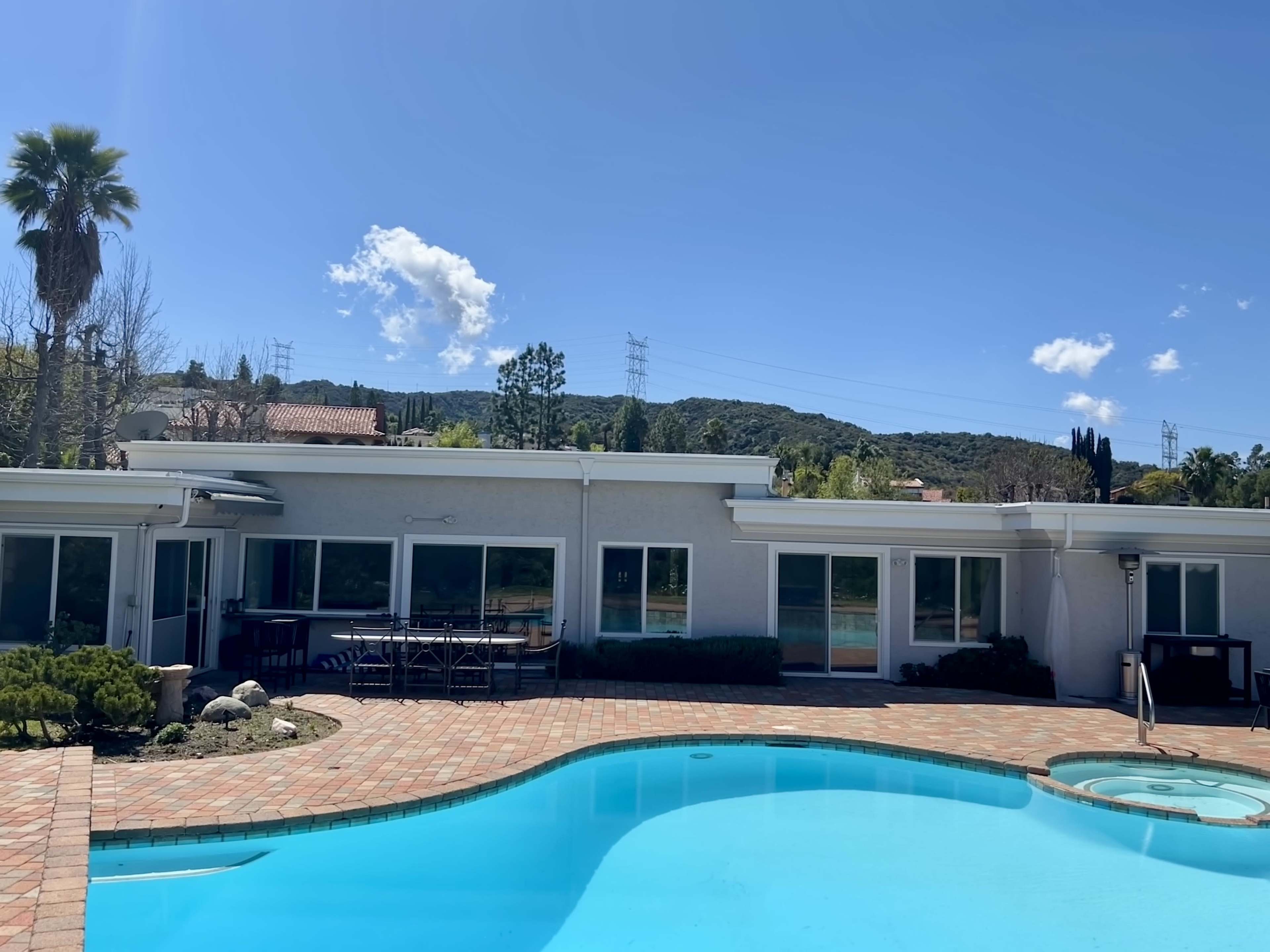 The image shows a swimming pool with a spa surrounded by a patio, adjacent to a single-story building and set against a backdrop of hills and a clear blue sky.