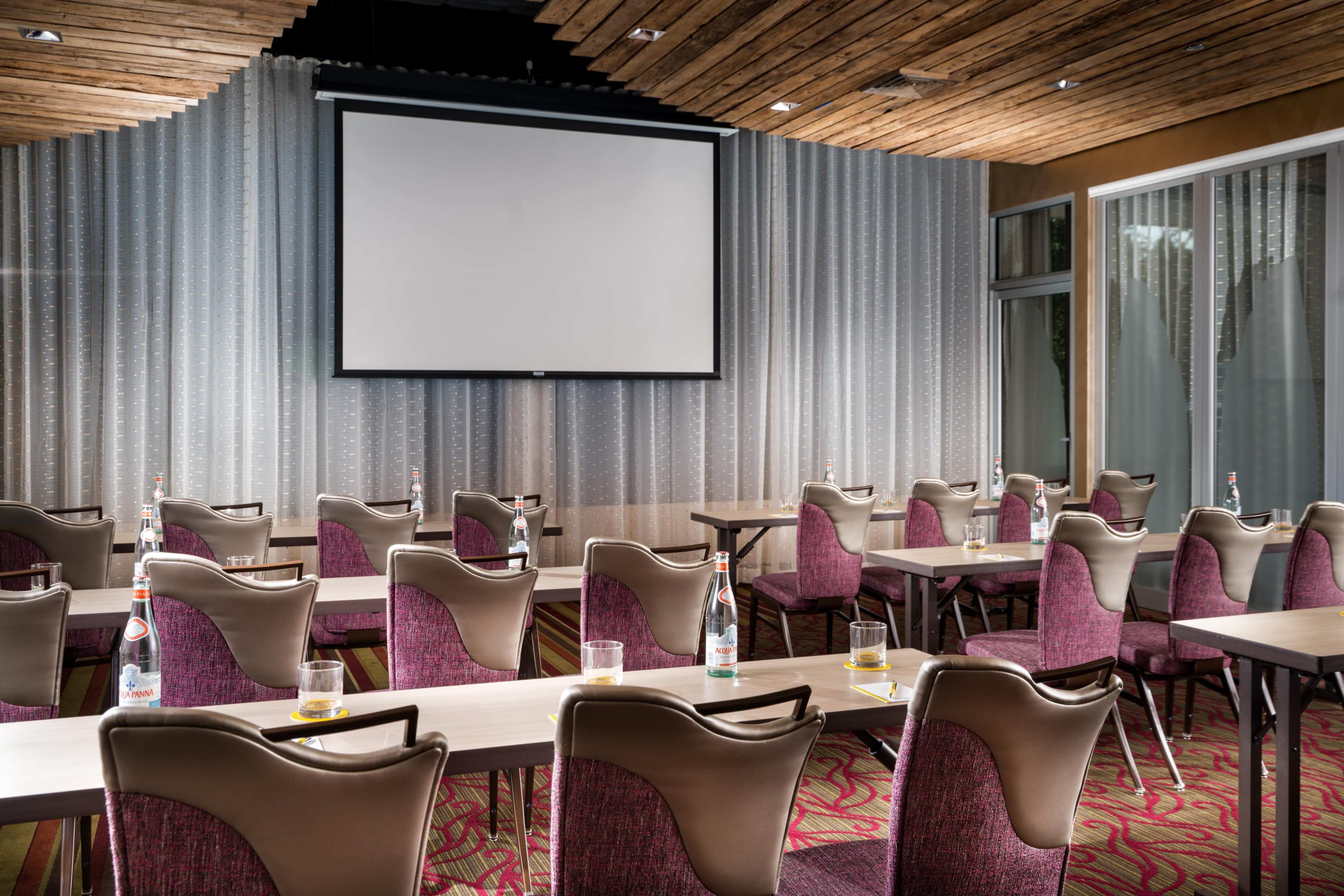 A conference room is set up with rows of tables and chairs facing a large projection screen, with bottled water and glasses on each table.