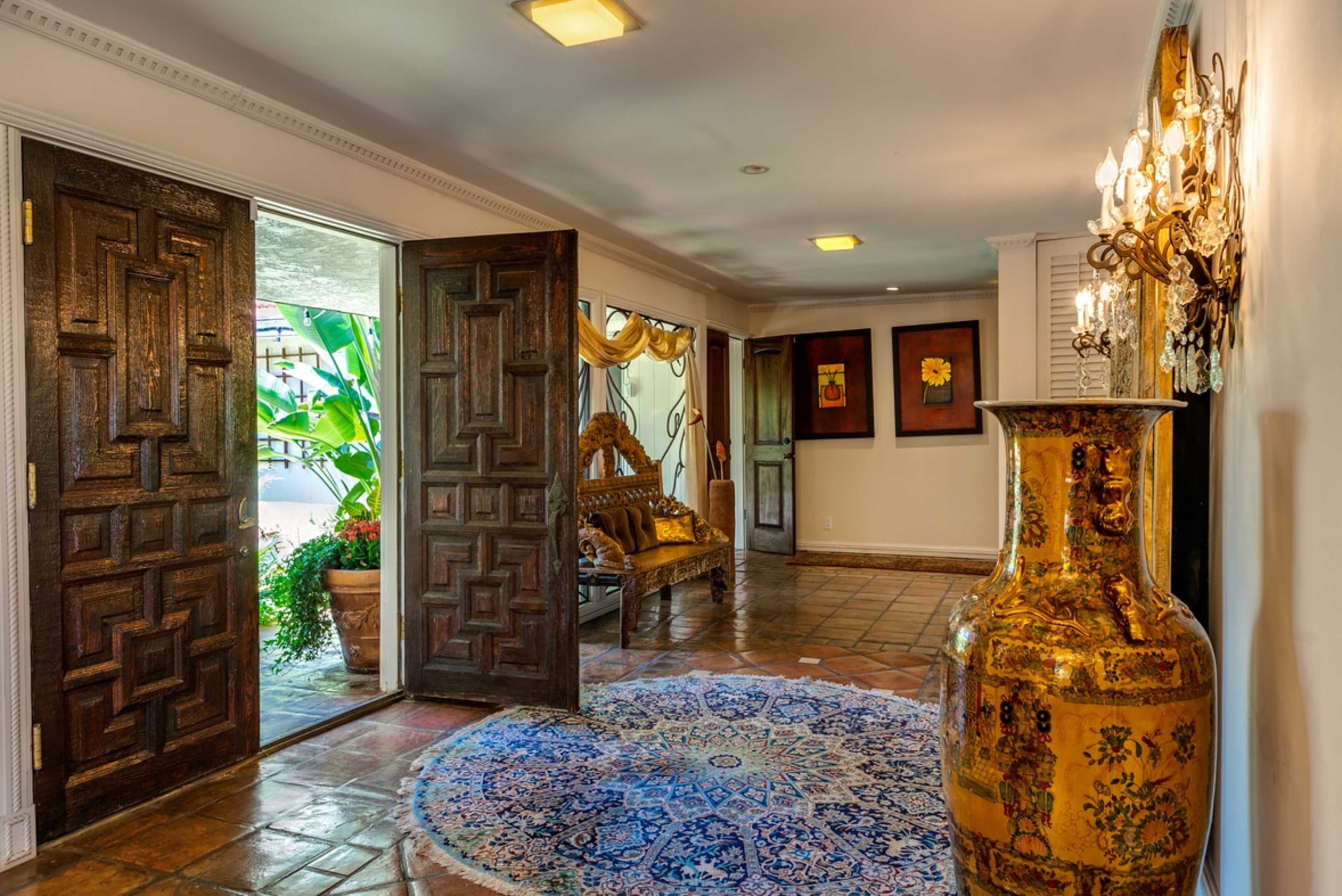 A spacious entryway features large wooden doors, a decorative vase, a circular rug, and a bench with elaborate carvings, complemented by wall art and indoor plants.