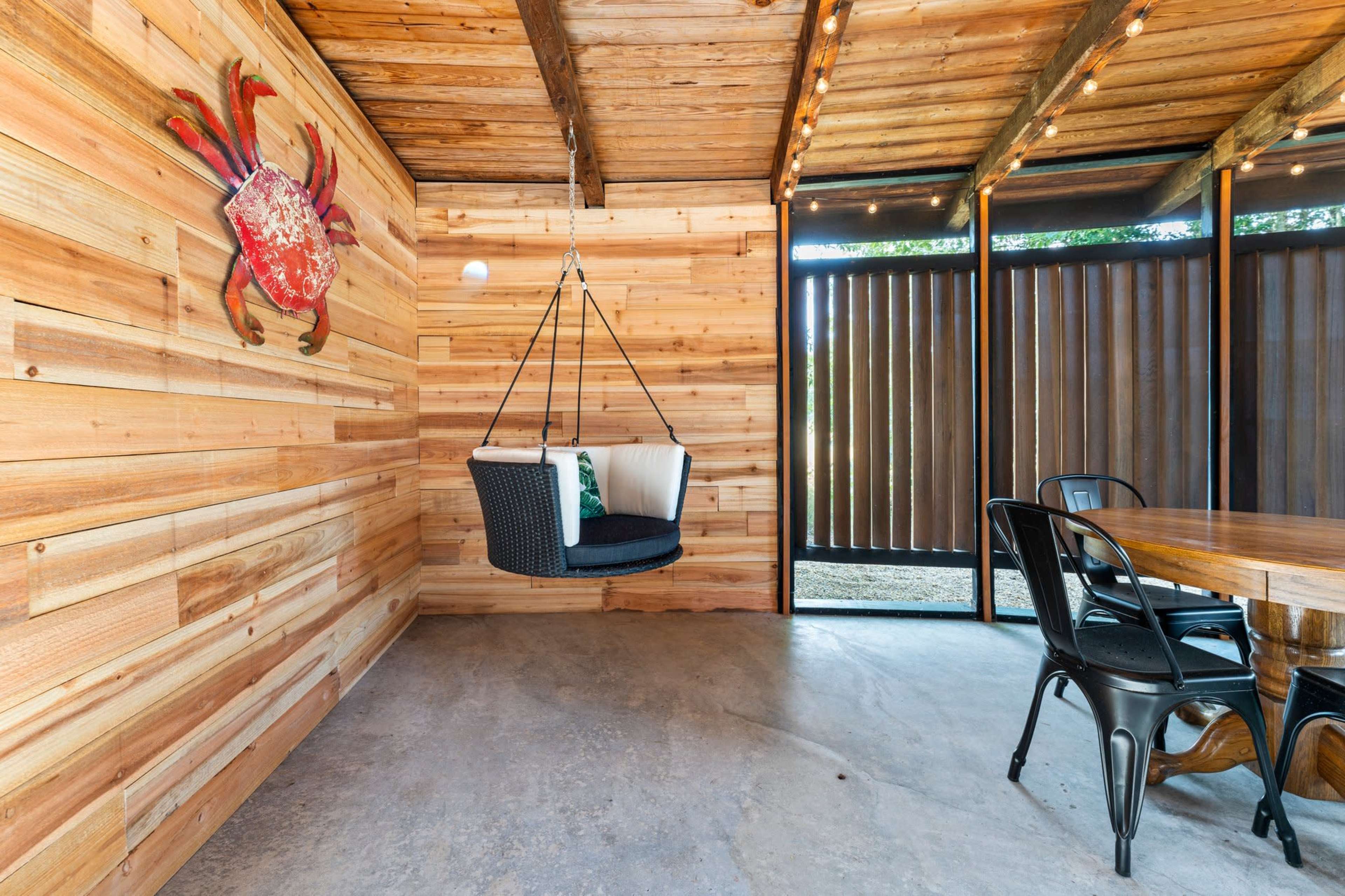 The image shows a wooden interior space featuring a swing chair, a dining table with black metal chairs, and a crab decoration on the wall.