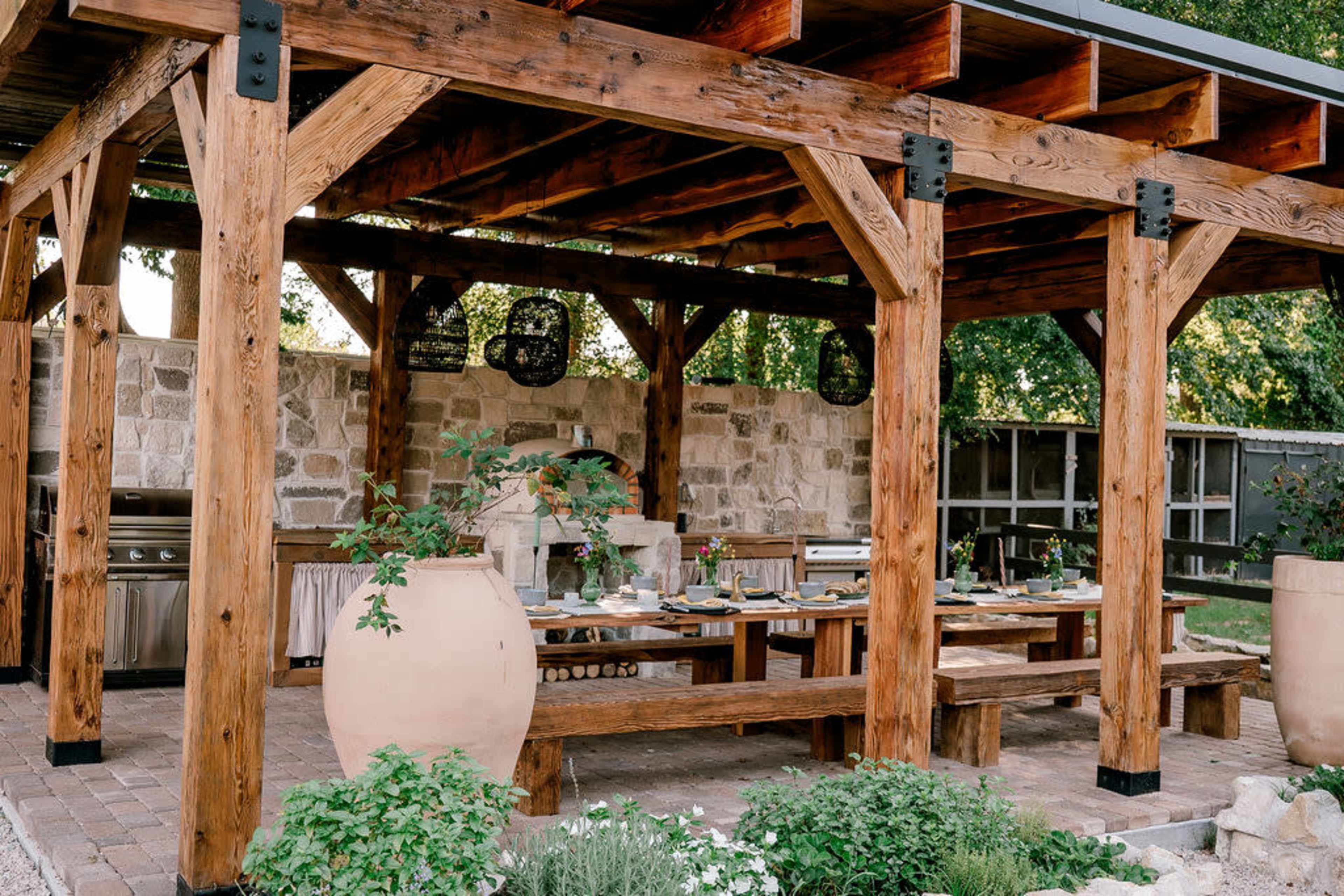 The image shows a wooden pavilion with a stone wall backdrop, featuring an outdoor kitchen area and a long dining table set for a meal.