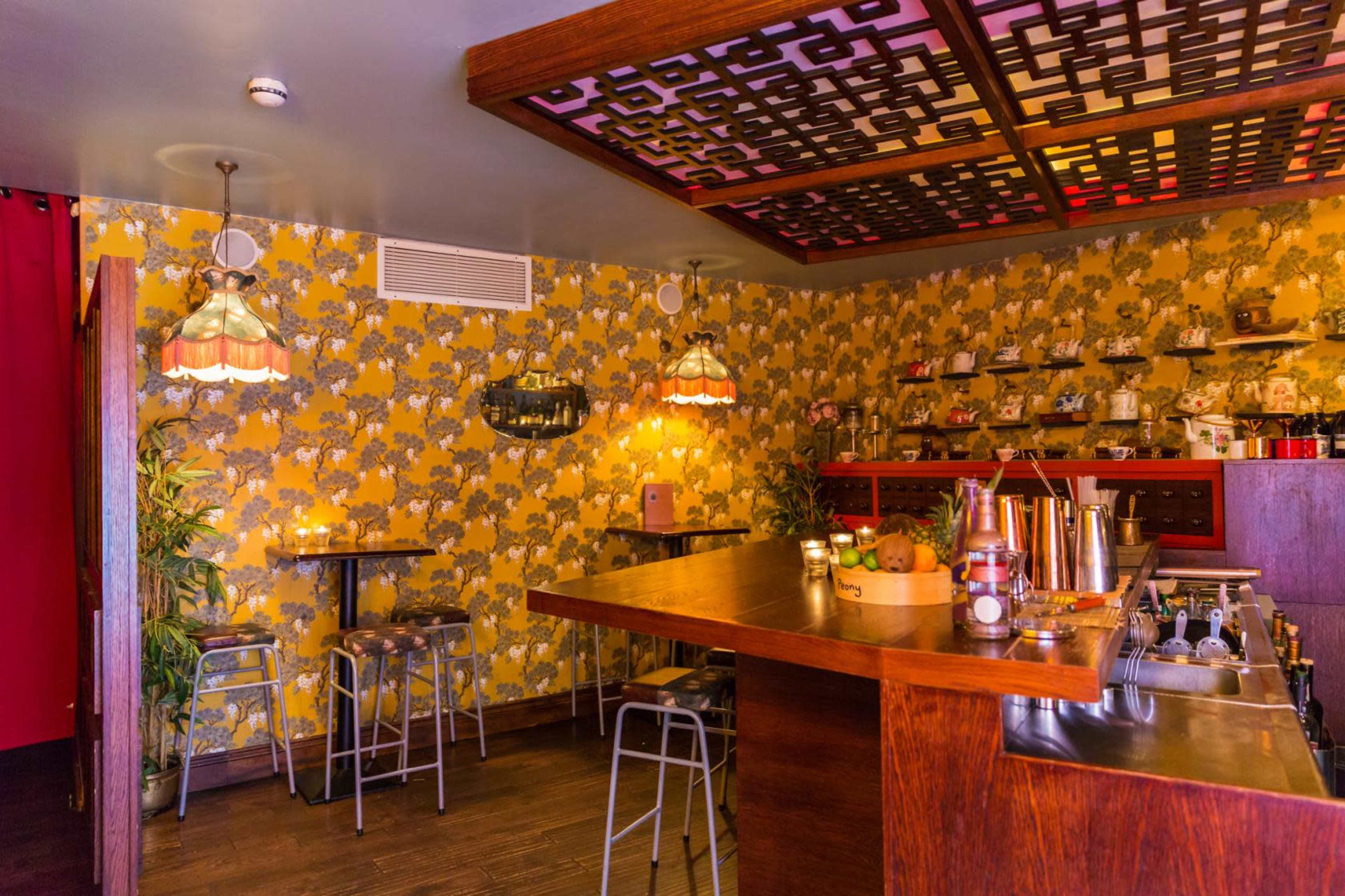 The image depicts a cozy bar area with wooden counters, metal stools, and decorative floral wallpaper.
