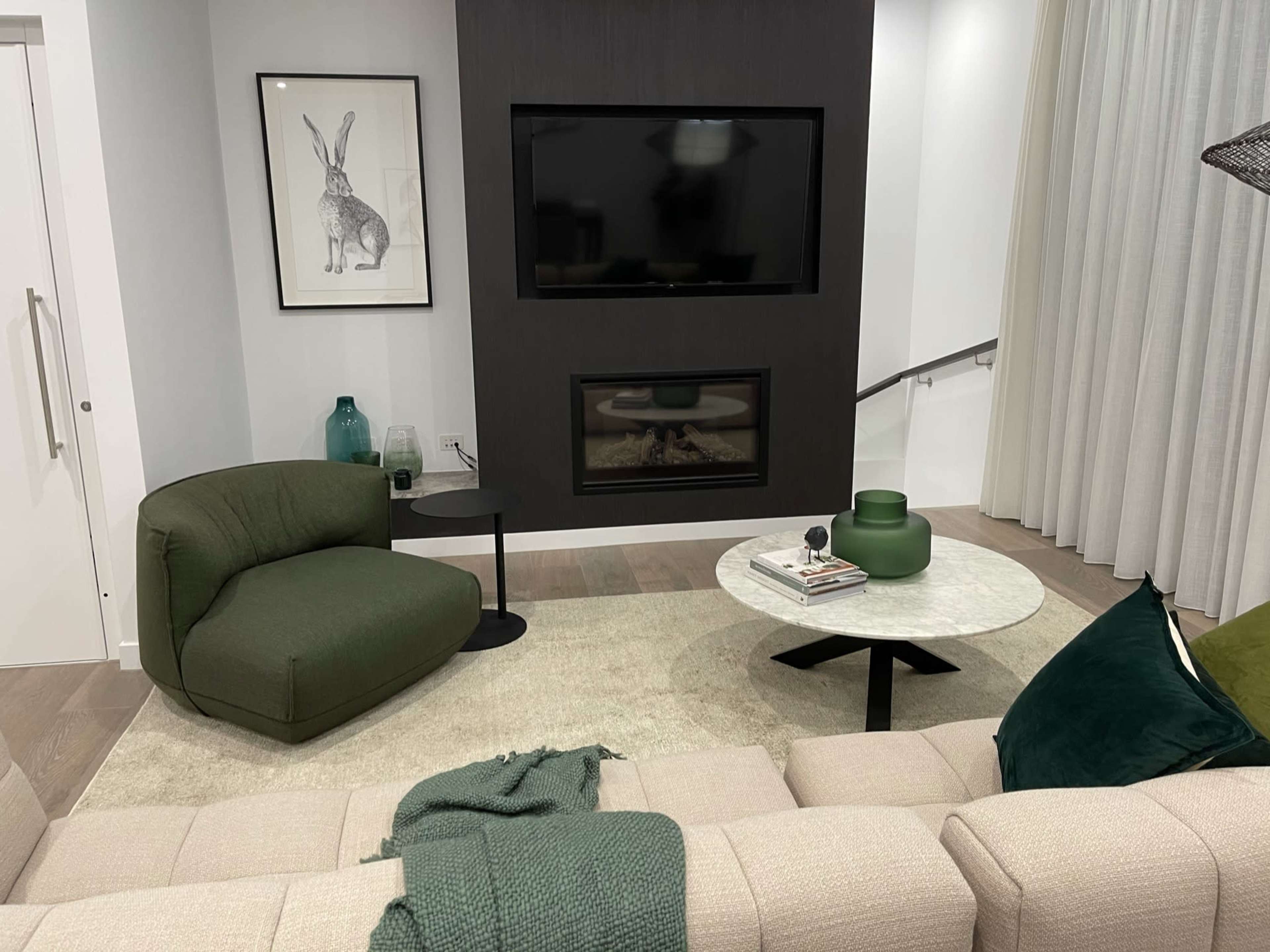 A modern living room features a green upholstered chair, a marble coffee table, and a wall-mounted television above a fireplace.