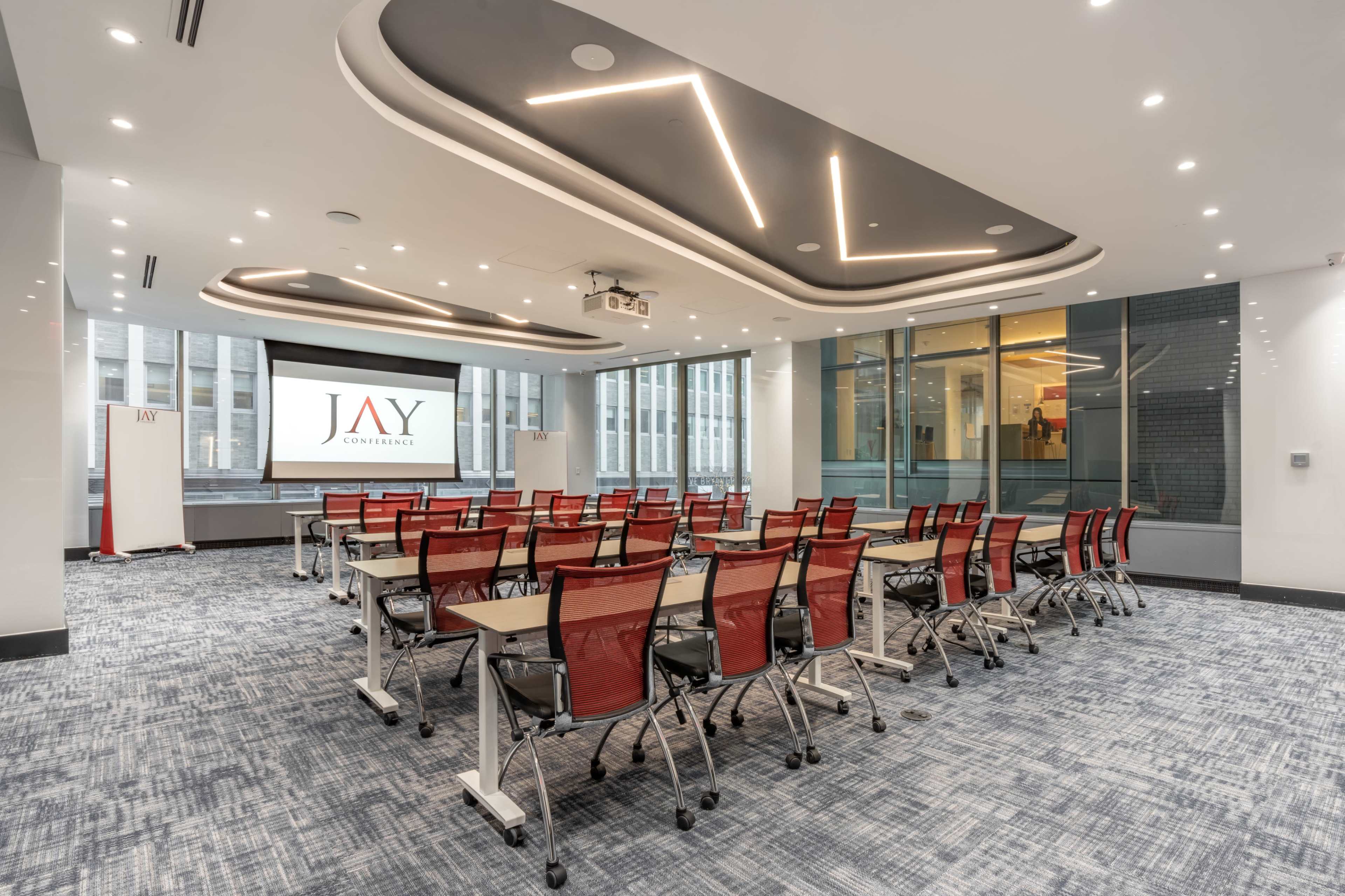 A modern conference room set up with rows of red mesh chairs facing a large screen displaying the logo "JAY" and a spacious layout with windows.