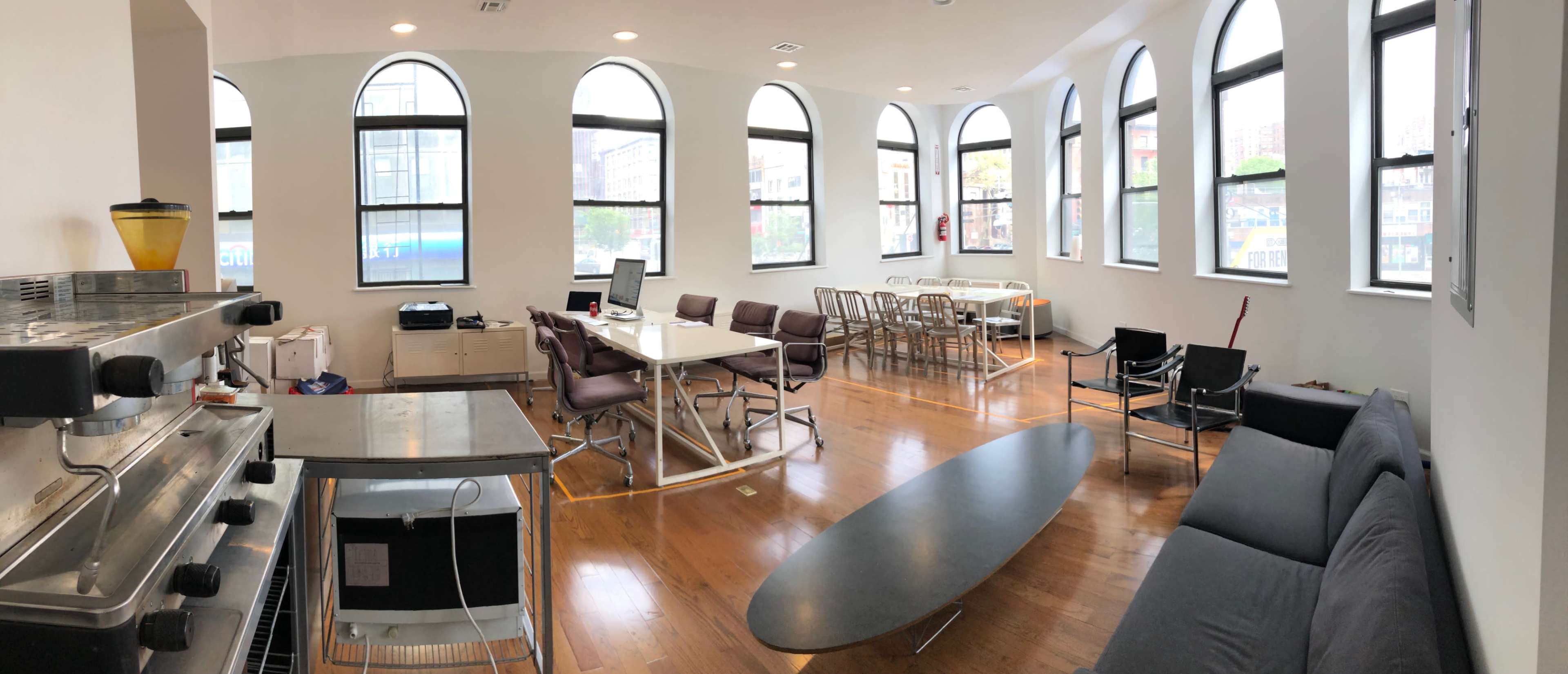 The image shows a modern, open-plan office space with large arched windows, a coffee machine, seating areas, and multiple tables arranged for work and meetings.