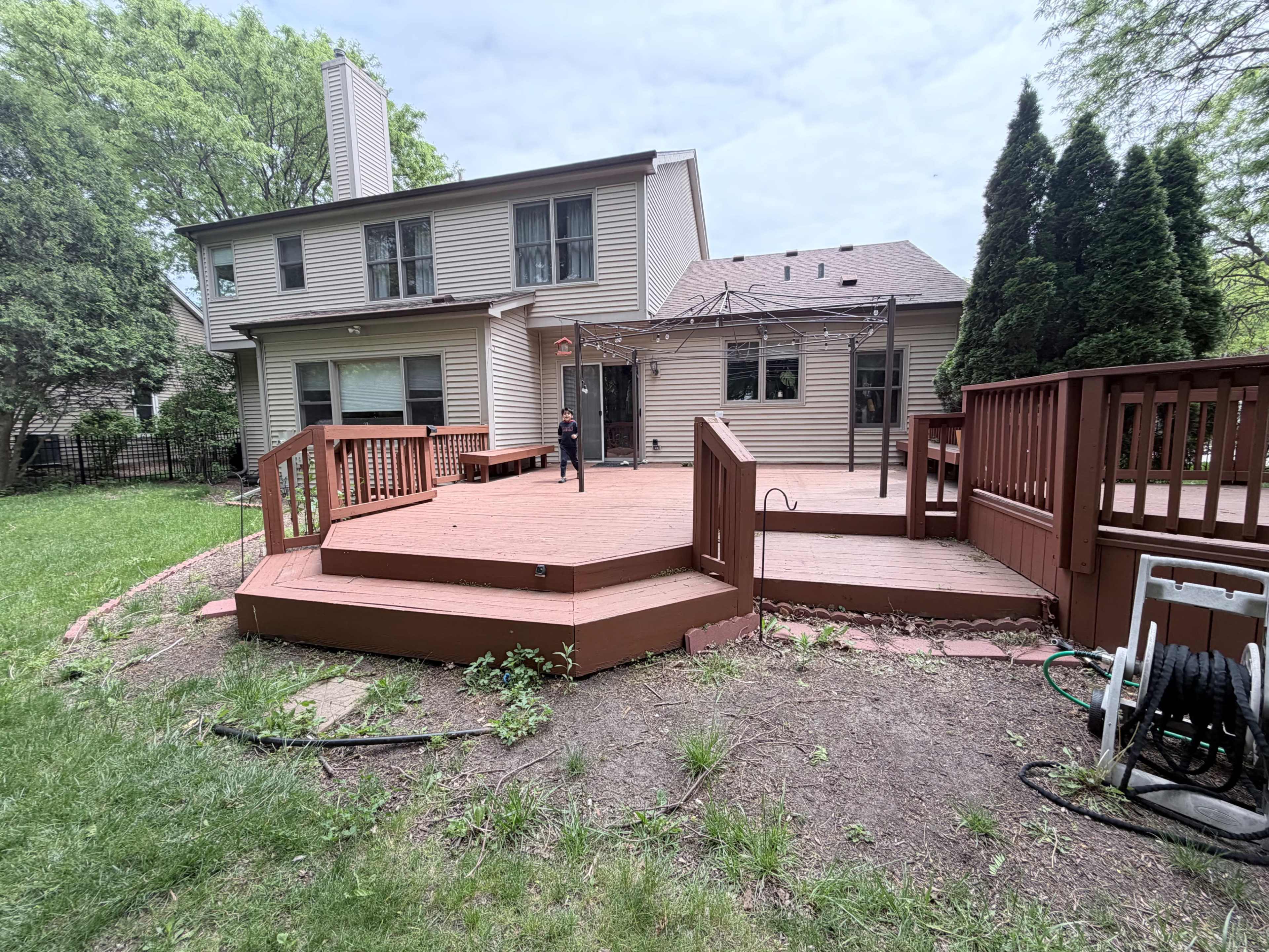A person stands on a wooden deck in the backyard of a two-story house surrounded by trees and grassy areas.