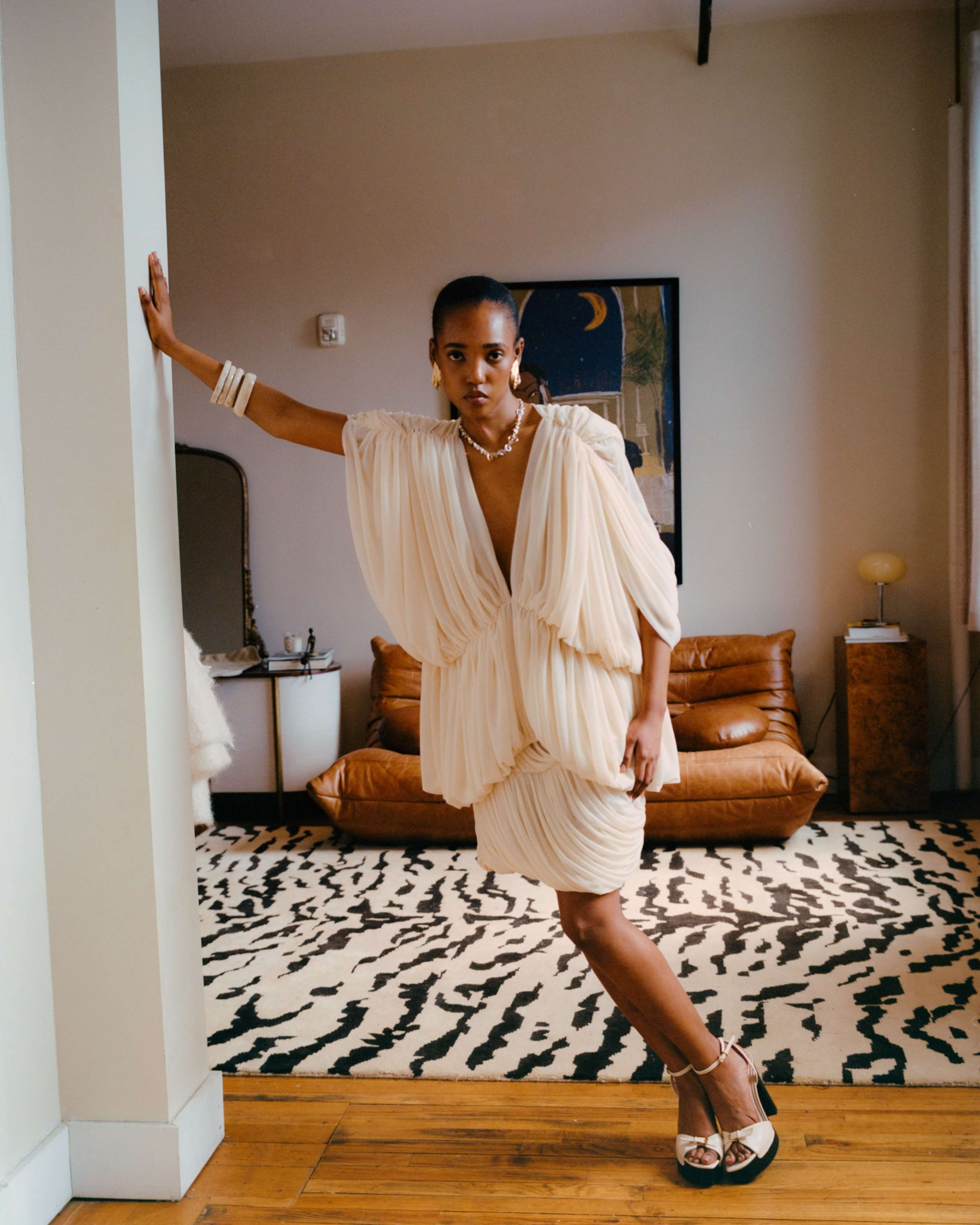 A person is striking a pose in a draped cream-colored dress while standing in a room with a patterned rug and mid-century furniture.