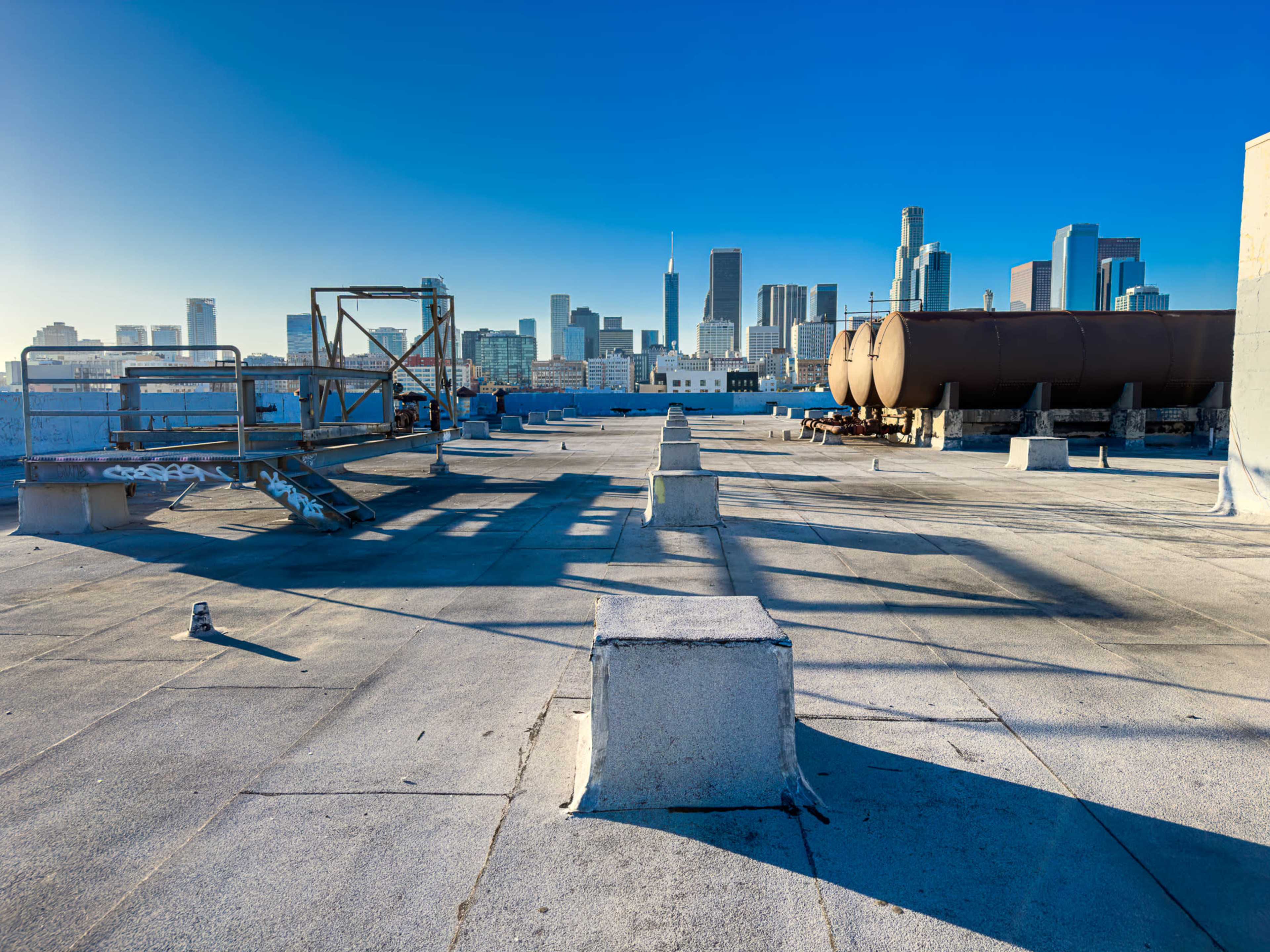 A rooftop area in an urban setting features several concrete blocks and large cylindrical tanks with a skyline of tall buildings in the background.