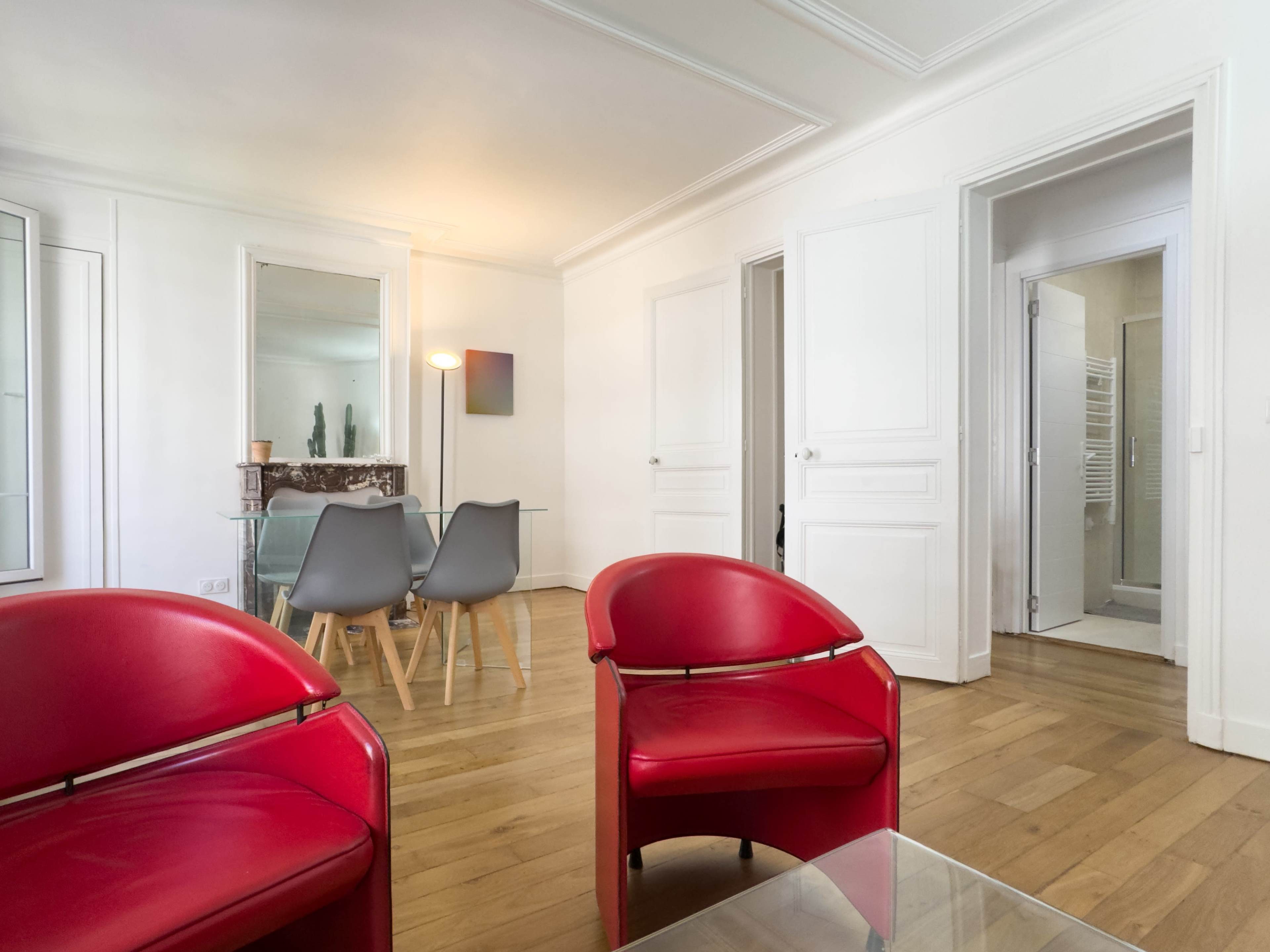 The image shows a modern living room with red chairs, wooden flooring, and adjacent doorways leading to other rooms.