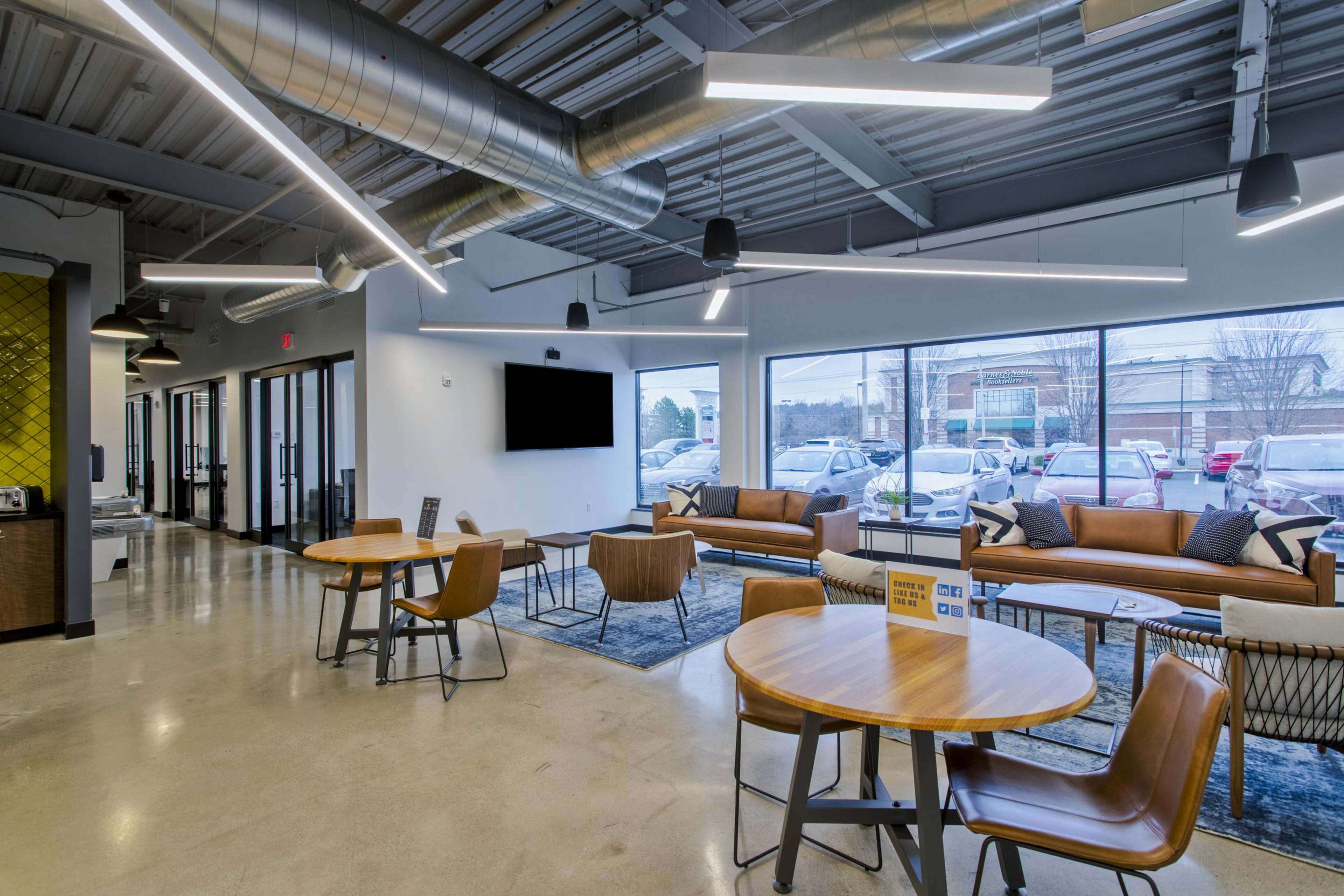 A modern lounge area featuring brown leather seating, wooden tables, and large windows providing natural light.