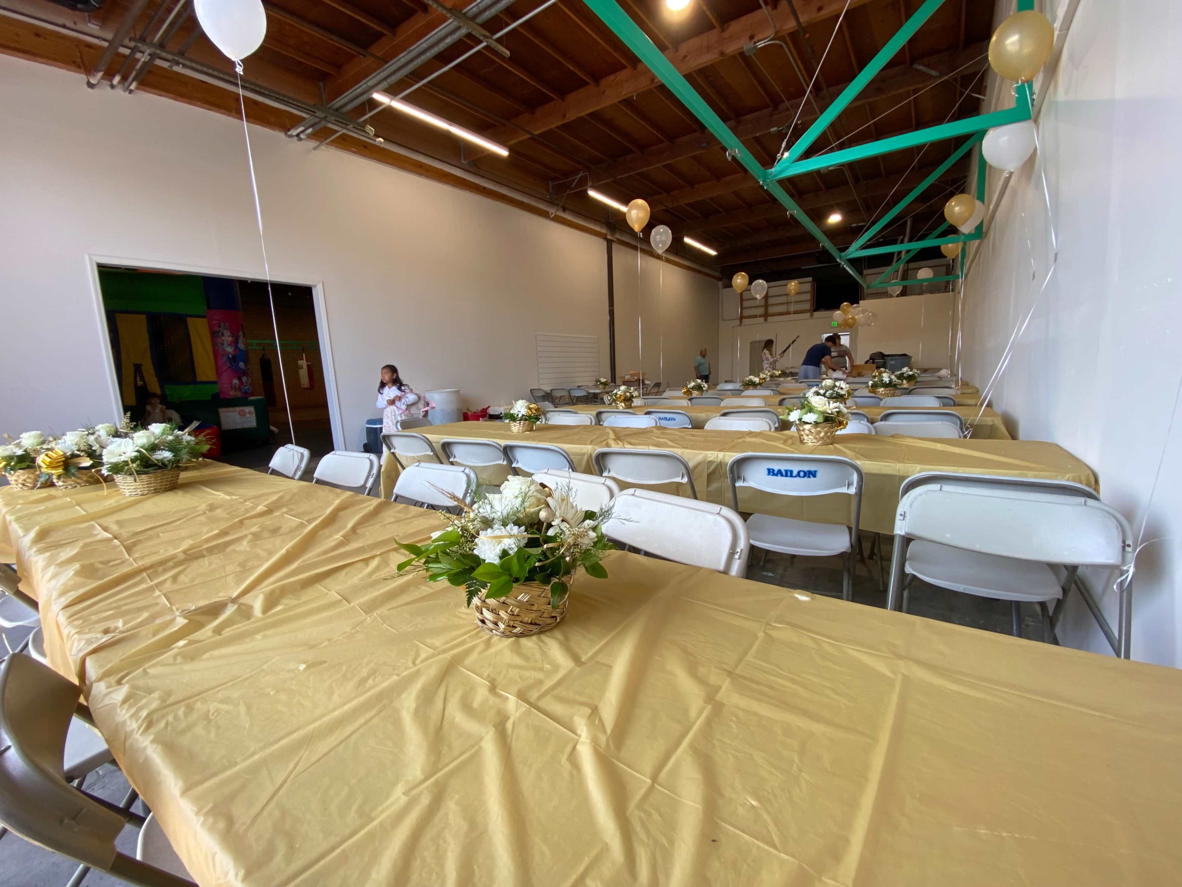 A neatly arranged event space with several long tables covered in gold tablecloths, each adorned with floral centerpieces, and empty chairs positioned around them.
