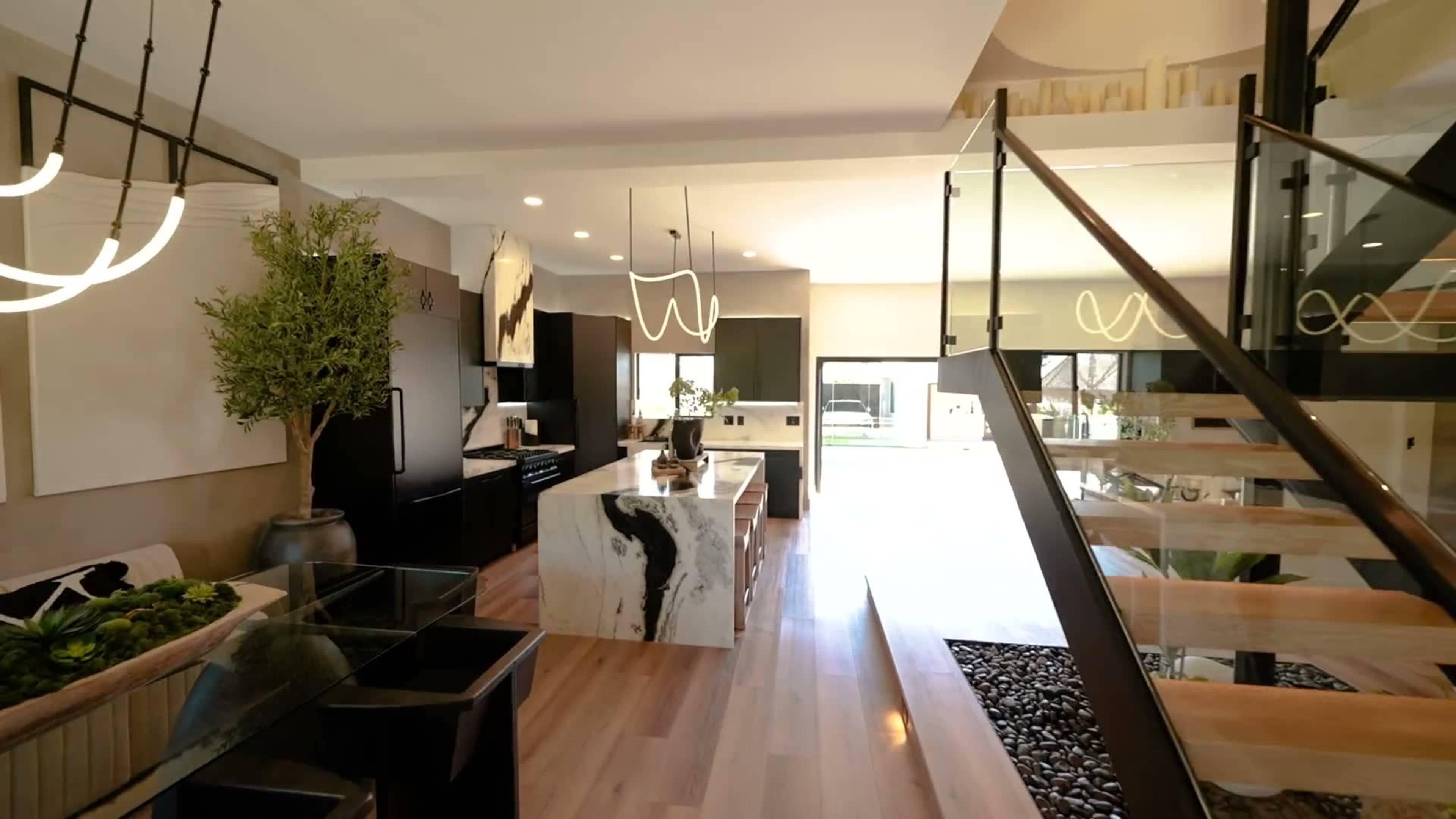 The image shows a modern kitchen and dining area featuring a large marble island, dark cabinetry, and a staircase with a glass railing.