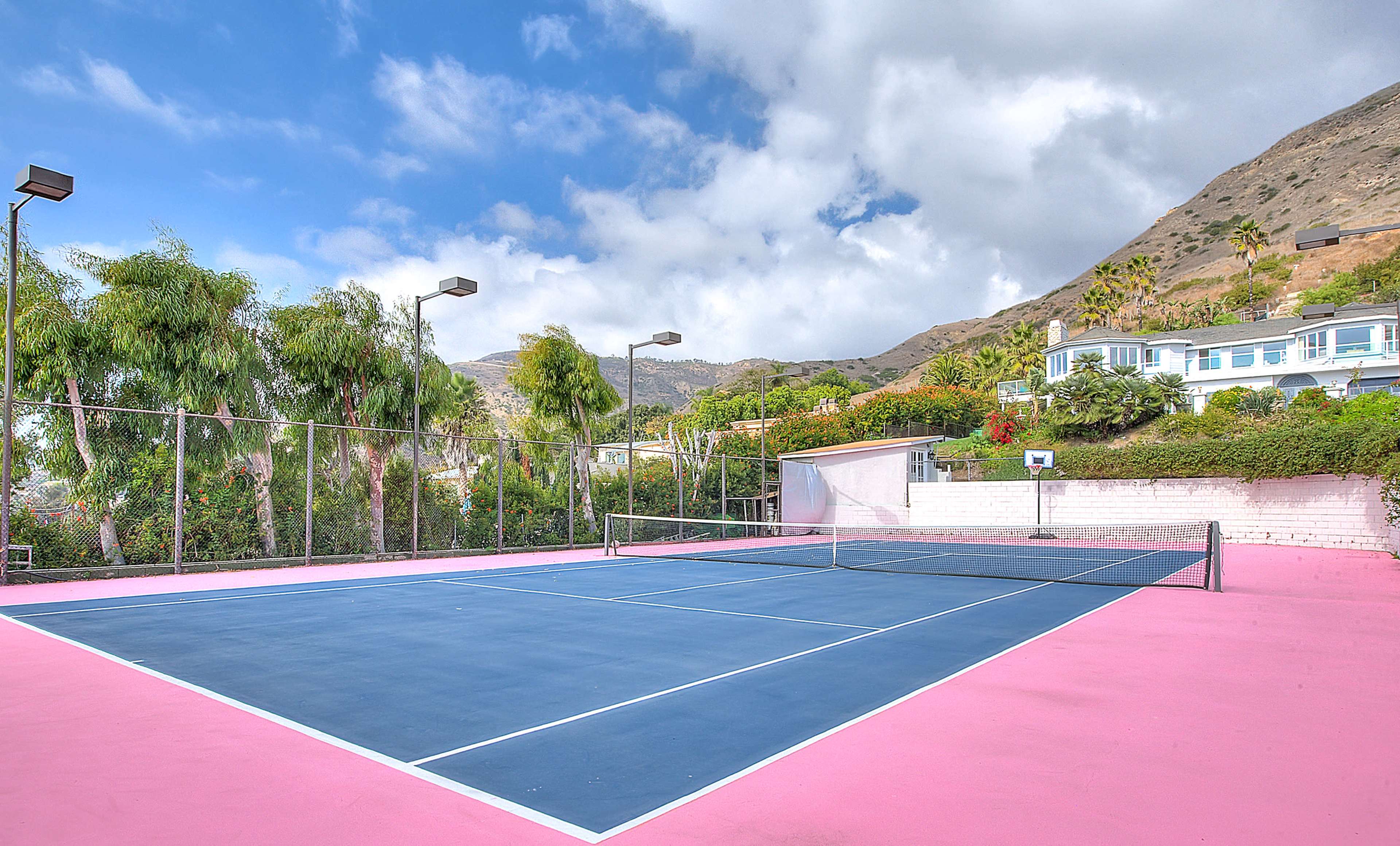 A tennis court with a blue playing surface surrounded by greenery and hills in the background.