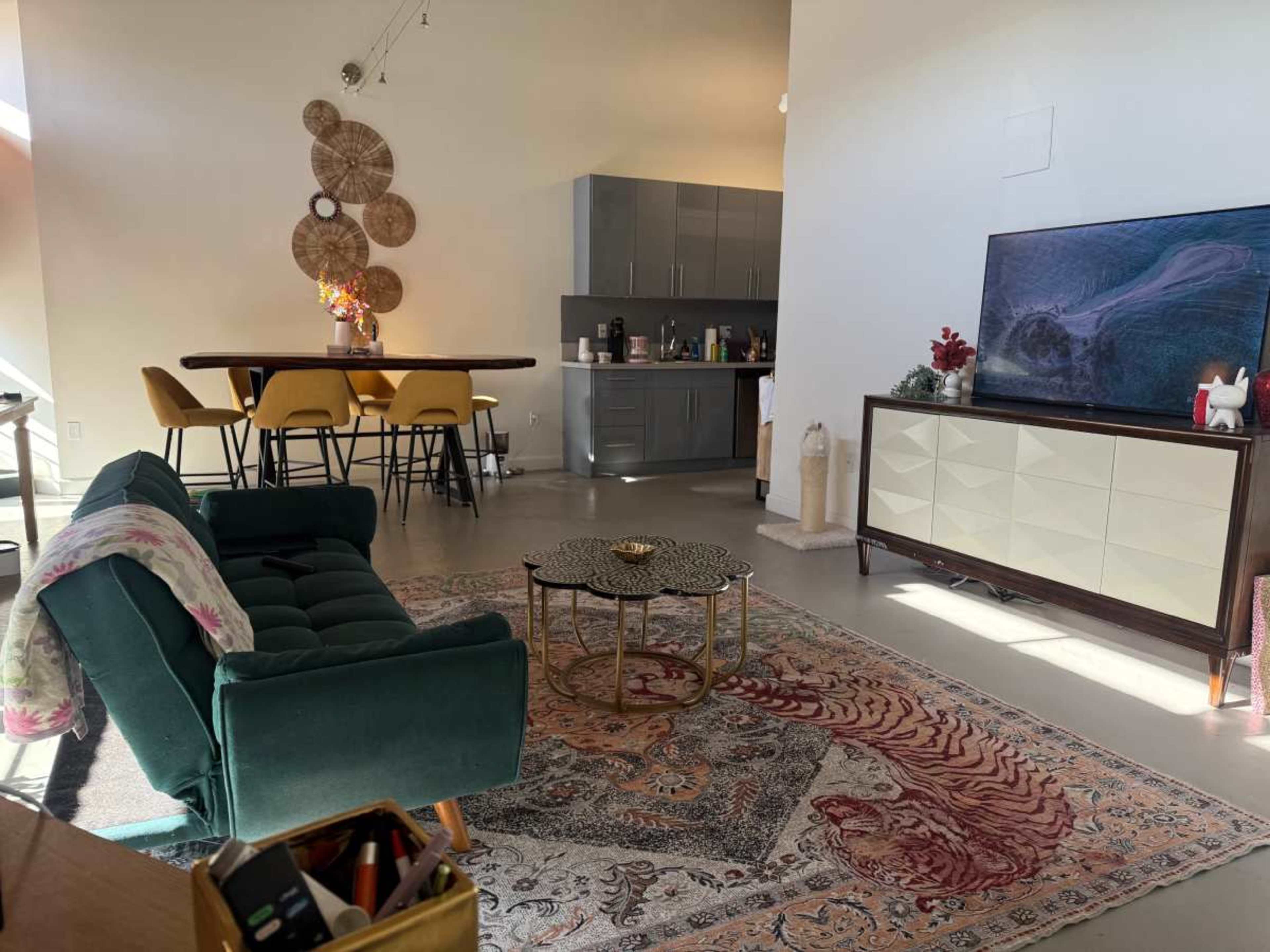 The image shows a modern living space featuring a green sofa, a coffee table, a TV stand, and a dining area with yellow chairs.