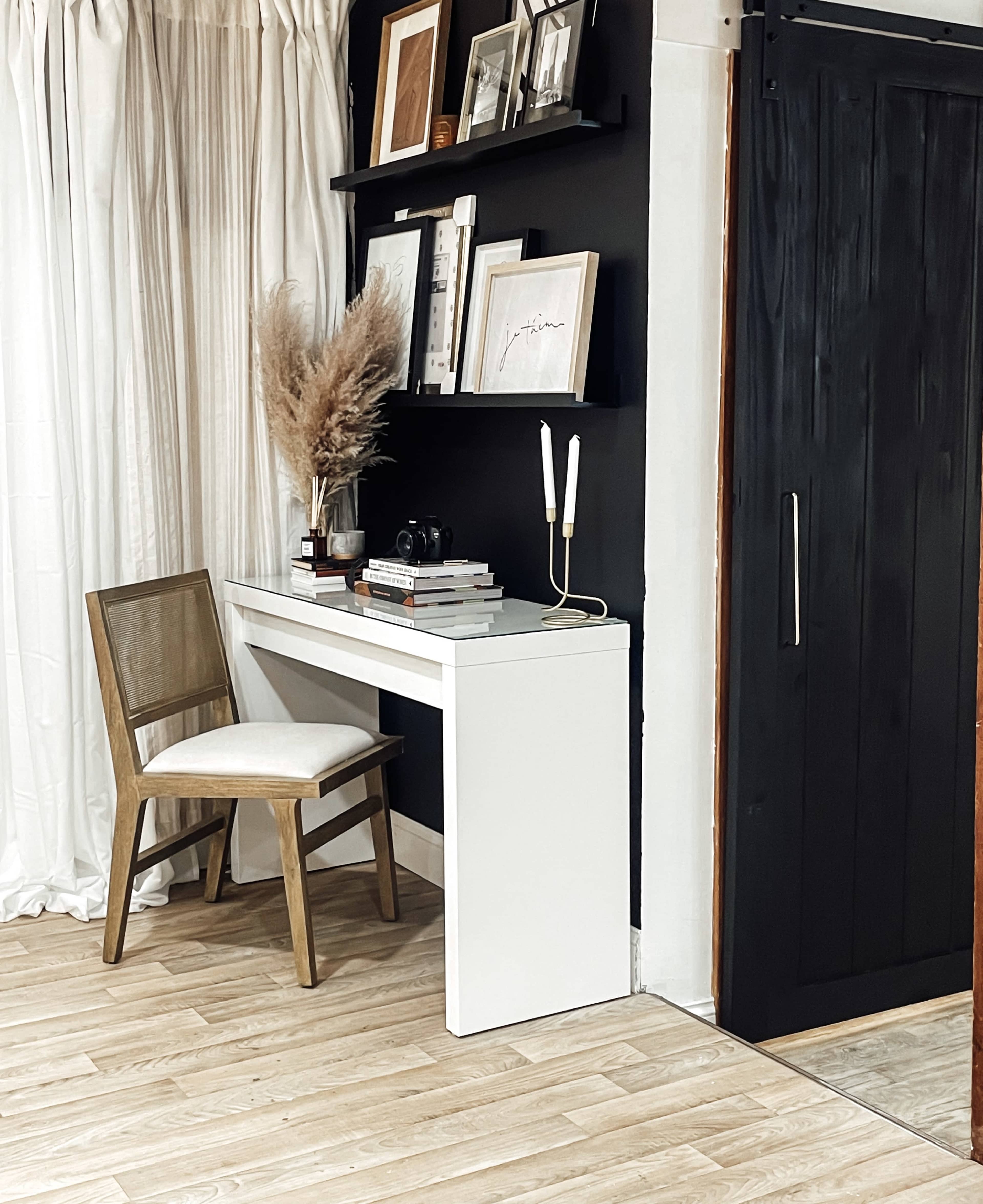 A minimalist workspace features a white desk with a chair, shelves holding framed art, and decor against a dark wall.