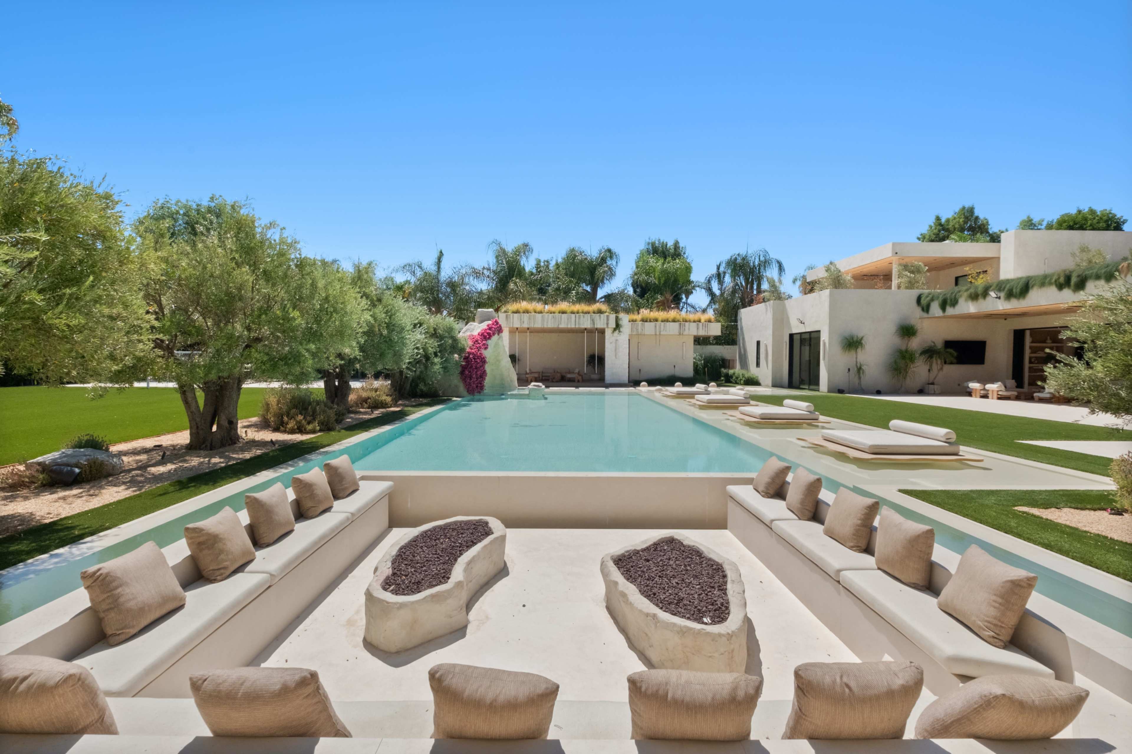 A modern outdoor lounge area with a sunken seating arrangement surrounds a rectangular swimming pool, framed by lush greenery and palm trees.